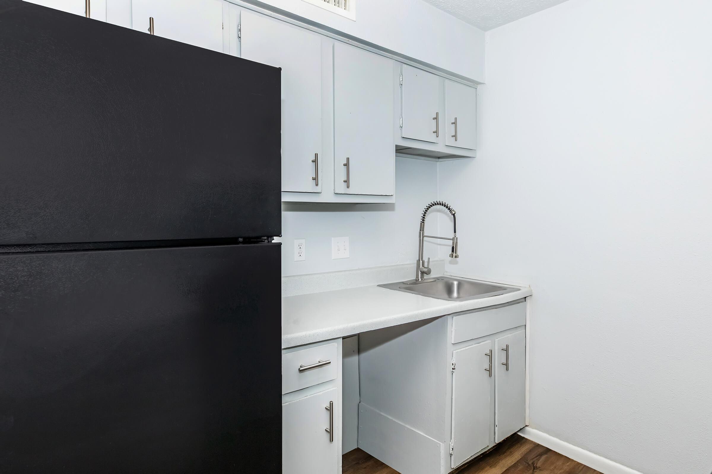 A modern kitchen featuring a black refrigerator, gray cabinets, and a stainless steel sink. The countertop is light, and the wall is painted in a soft white. The space is clean and minimalist, with wooden flooring.