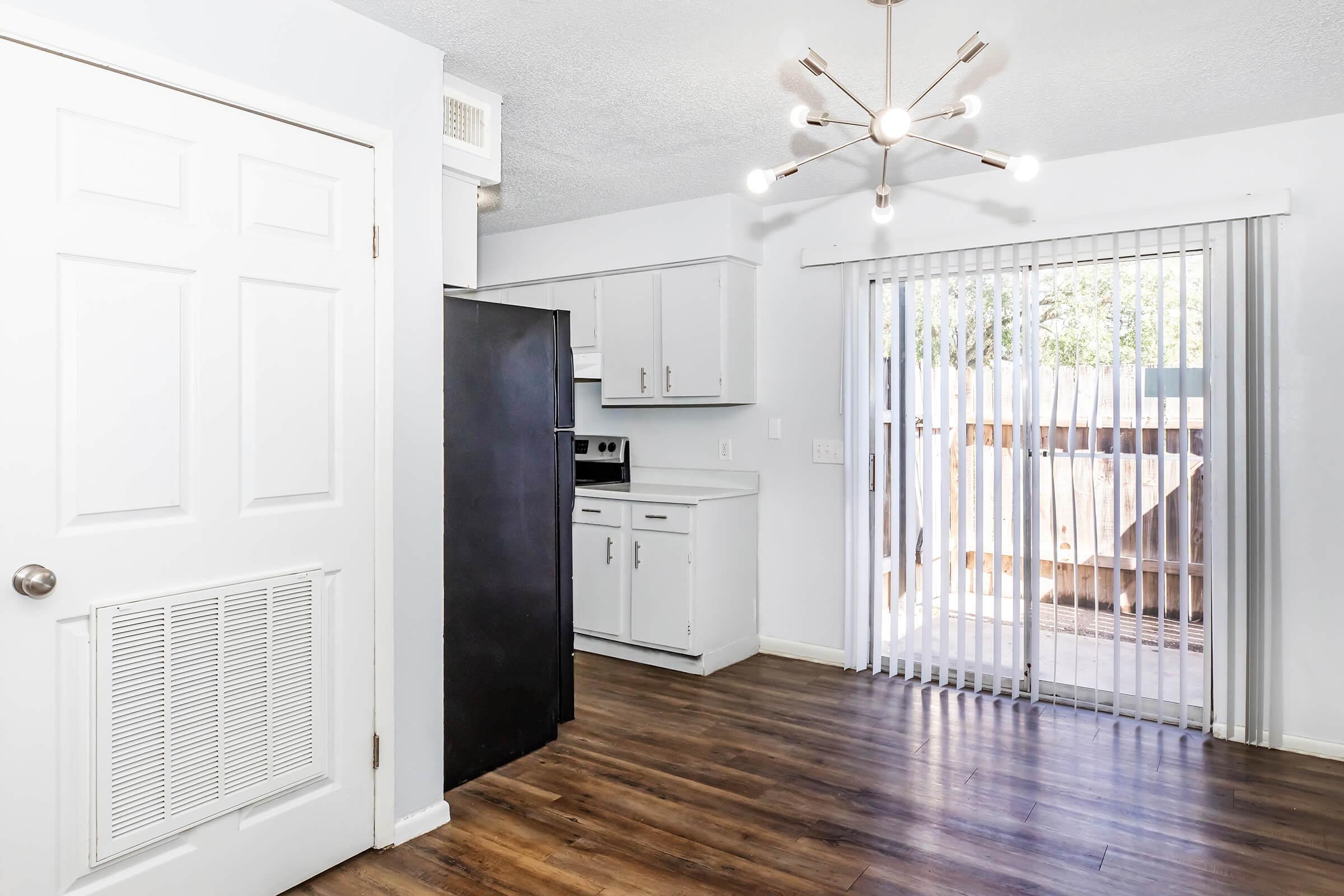 A modern kitchen featuring light-colored cabinets, a black refrigerator, and a sliding glass door leading to an outdoor area. The wooden floor adds warmth to the space, and soft lighting is provided by a contemporary chandelier. The room has a bright and inviting atmosphere.