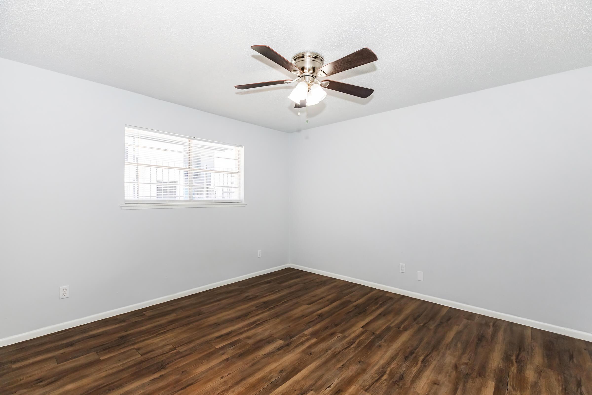 A spacious, empty room with light-colored walls and a wooden floor. The room features a ceiling fan with five blades, and there is a window letting in natural light. The overall atmosphere is bright and inviting, suitable for various uses.