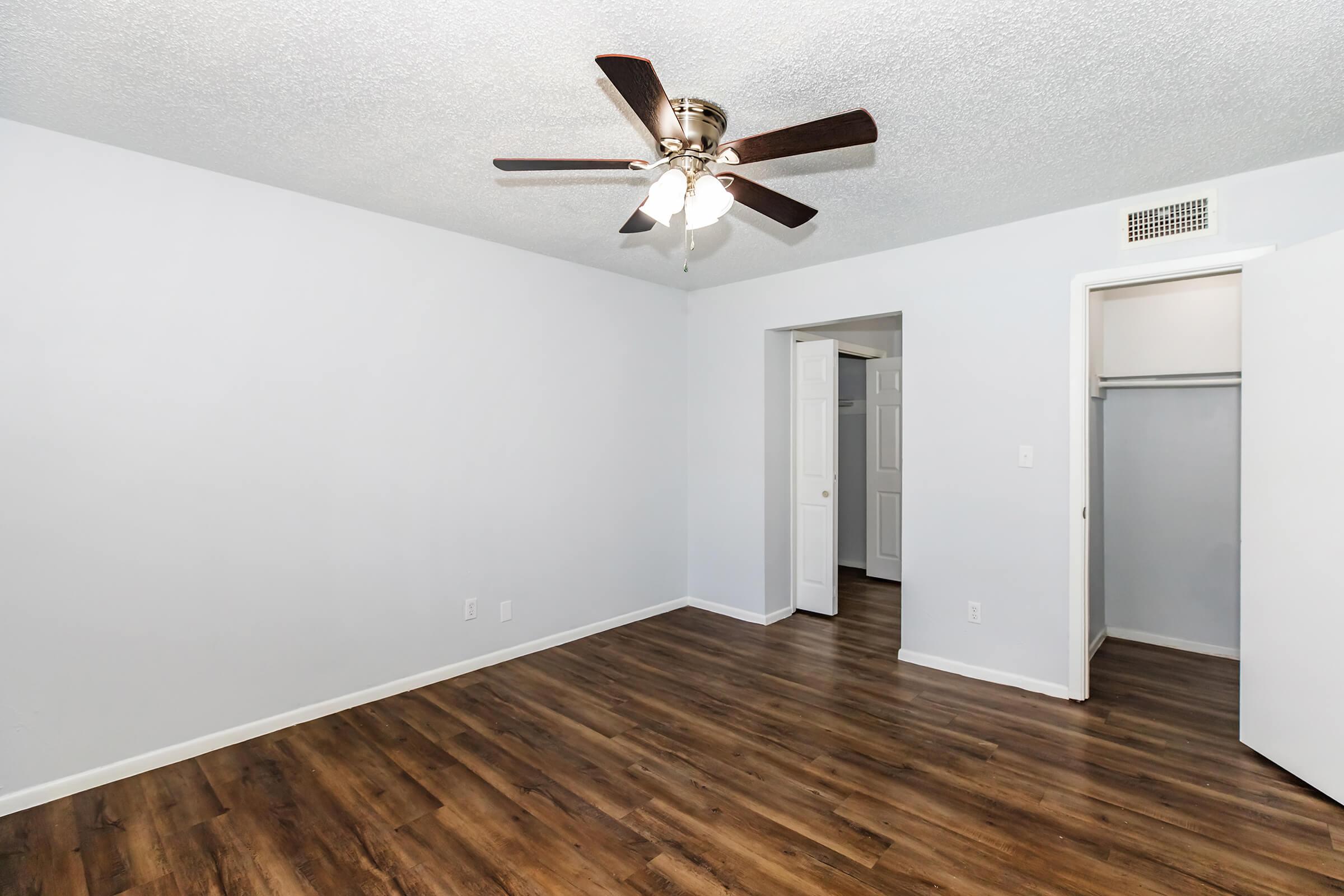 A spacious room with light blue walls and a ceiling fan. The floor is covered in wooden laminate. There are two doors—one leading to a closet and the other to an adjacent room. The space is well-lit, creating a welcoming atmosphere.