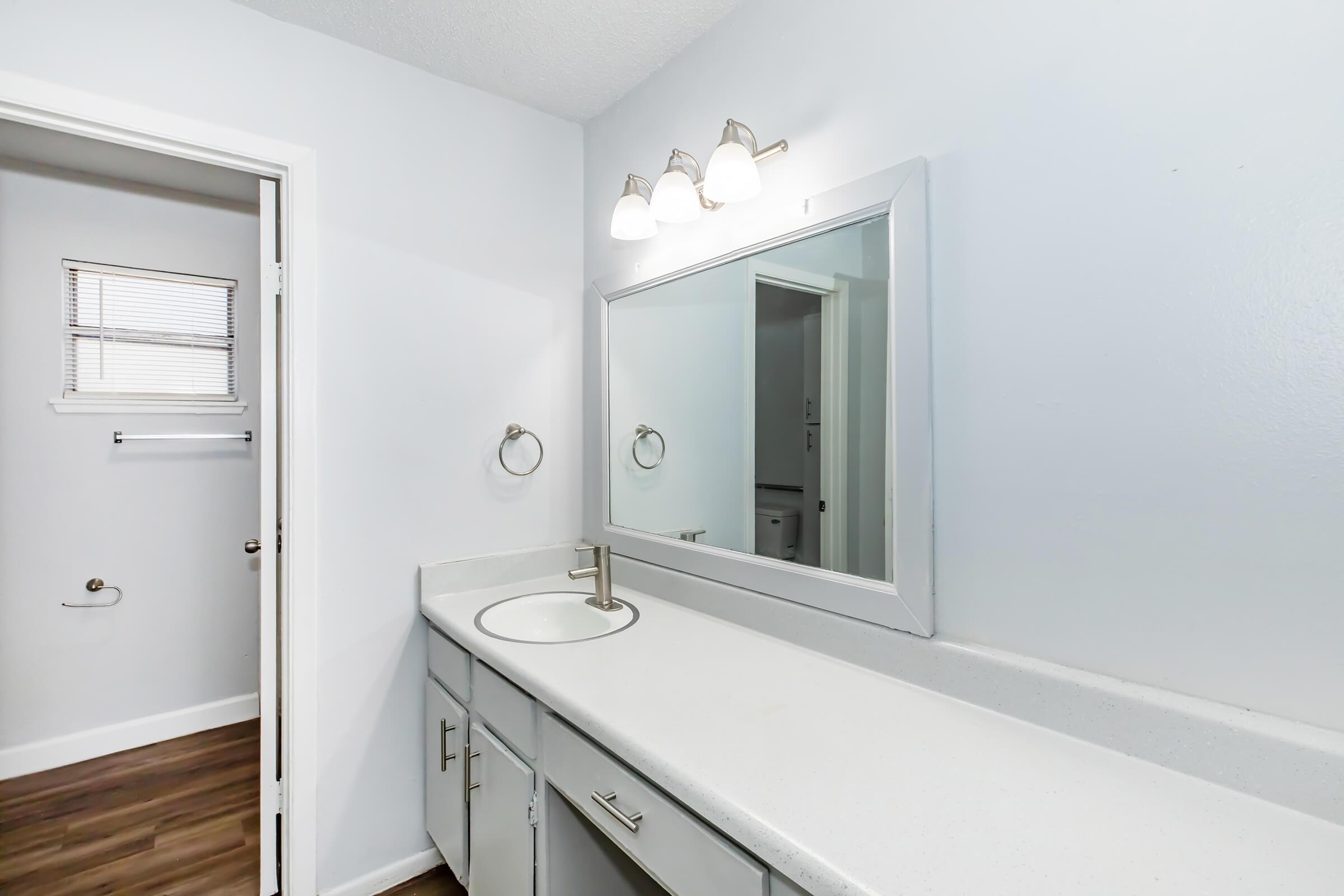 A clean, modern bathroom featuring a long white countertop with a sink, a large mirror above it, and three light fixtures. The walls are painted light blue, and there's a door leading to another room. A towel holder is mounted on the wall, and the flooring is a dark wood finish.