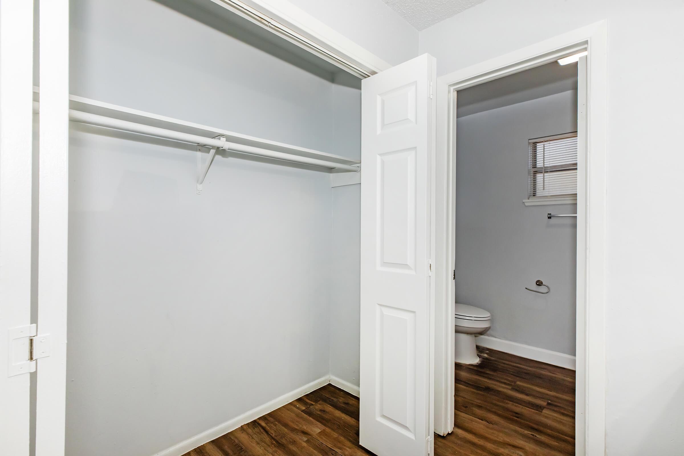Interior view of a small closet with a single hanging rod and light-colored walls. An open door leads to a bathroom area that includes a visible toilet. The floor features wooden planks, creating a modern and clean aesthetic.