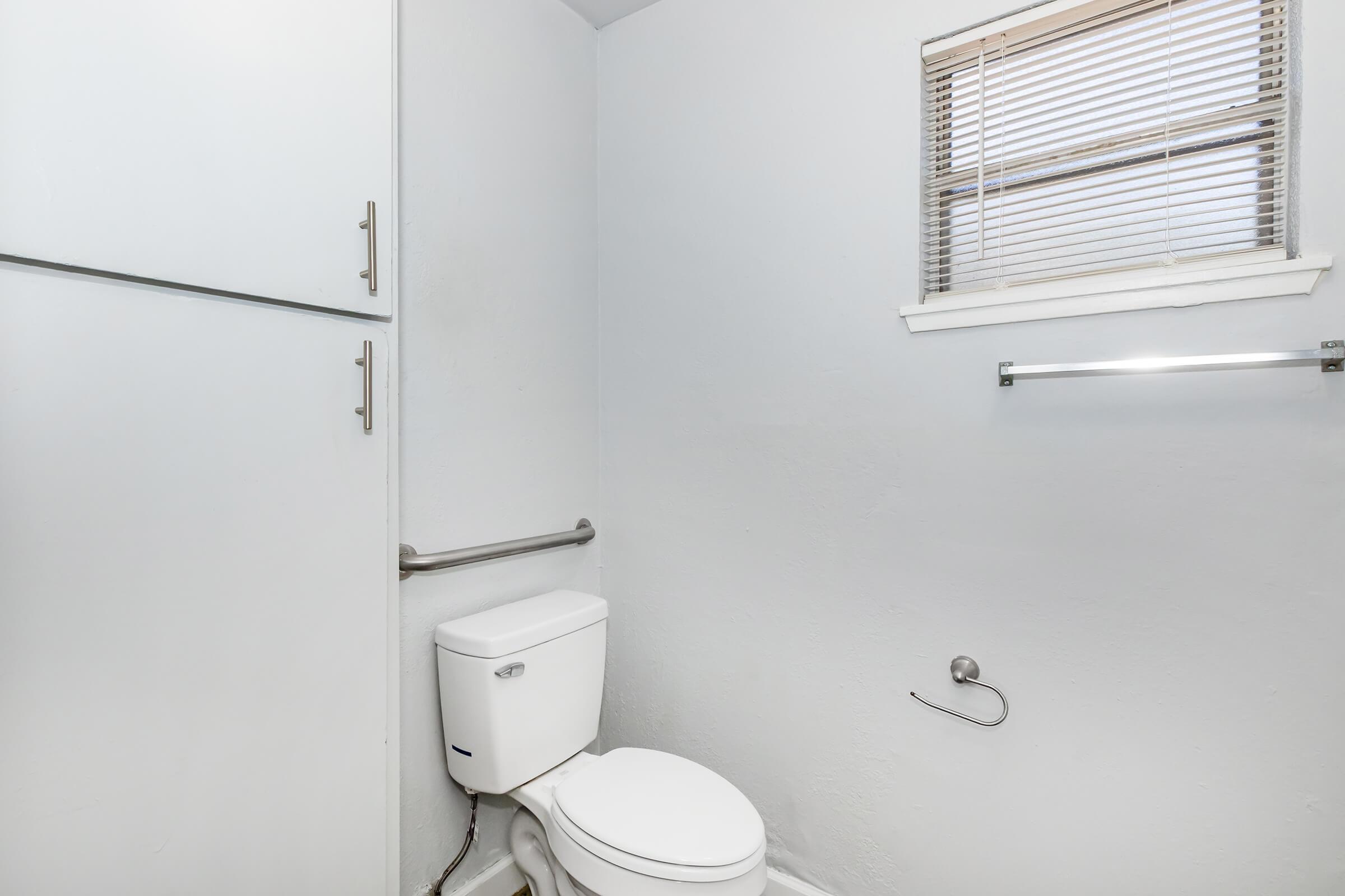 A simple bathroom with light gray walls. It features a white toilet, a small window covered with blinds, a towel rack, and a medicine cabinet. The overall design is minimalistic and clean, showcasing an uncluttered space.