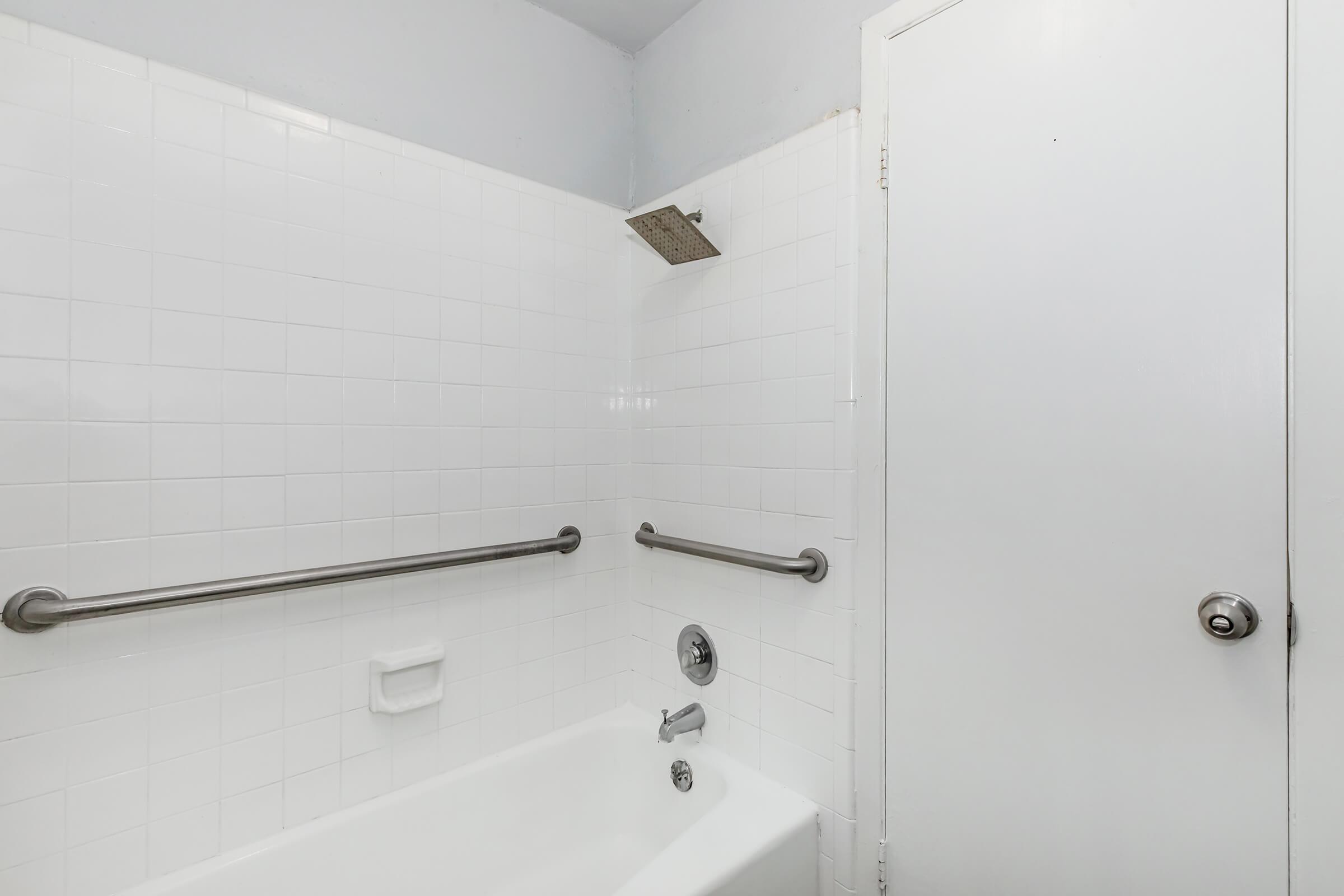 A clean, bright bathroom featuring a white tiled shower area with a built-in tub, a showerhead mounted on the wall, grab bars for safety, and a closed door on the right. The overall color scheme is minimalistic and modern.