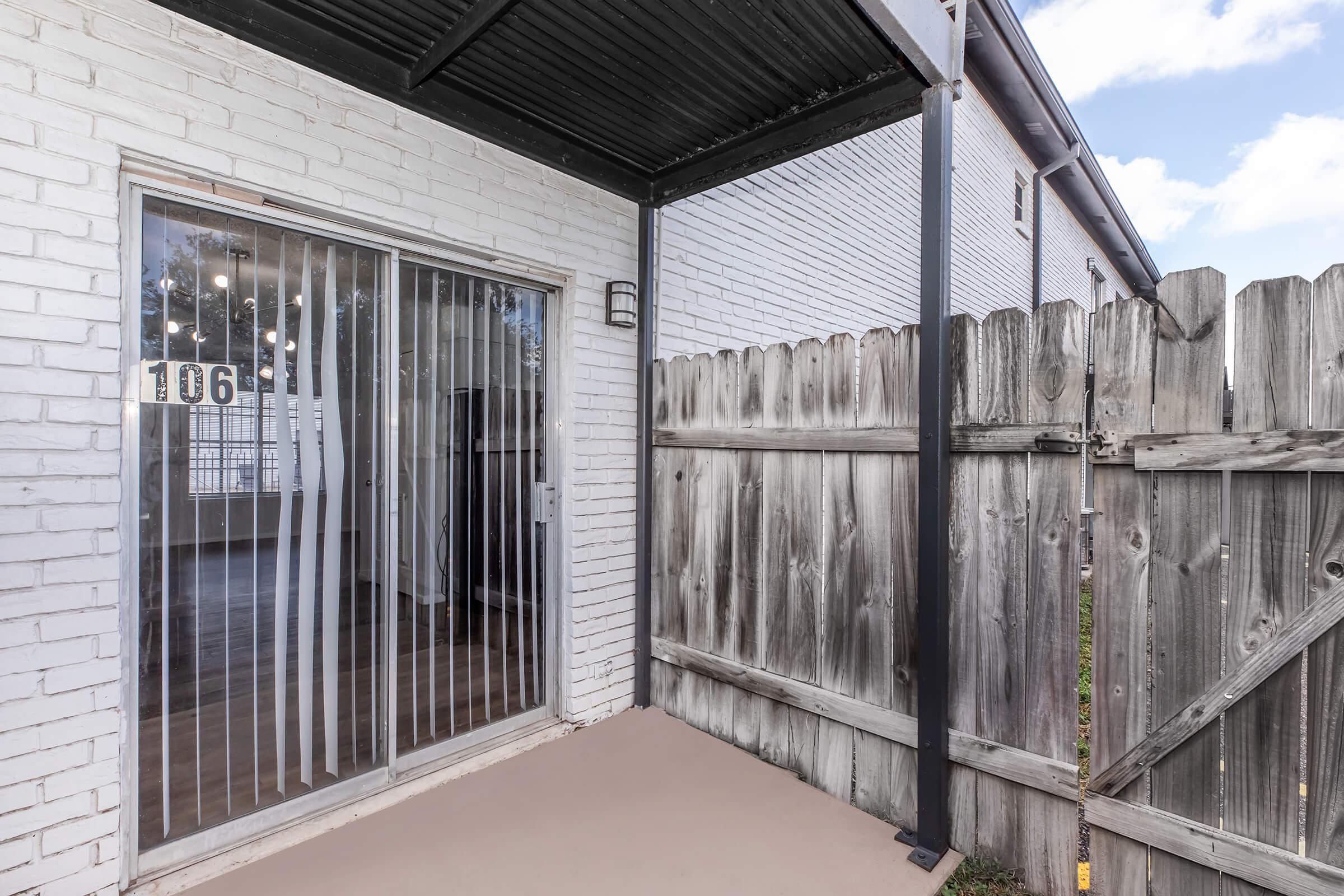 A view of a small outdoor patio area with a sliding glass door to a white-brick room. A wooden fence with a weathered appearance encloses the space, and the floor is a light-colored surface. Bright daylight is visible in the background, creating an inviting atmosphere.