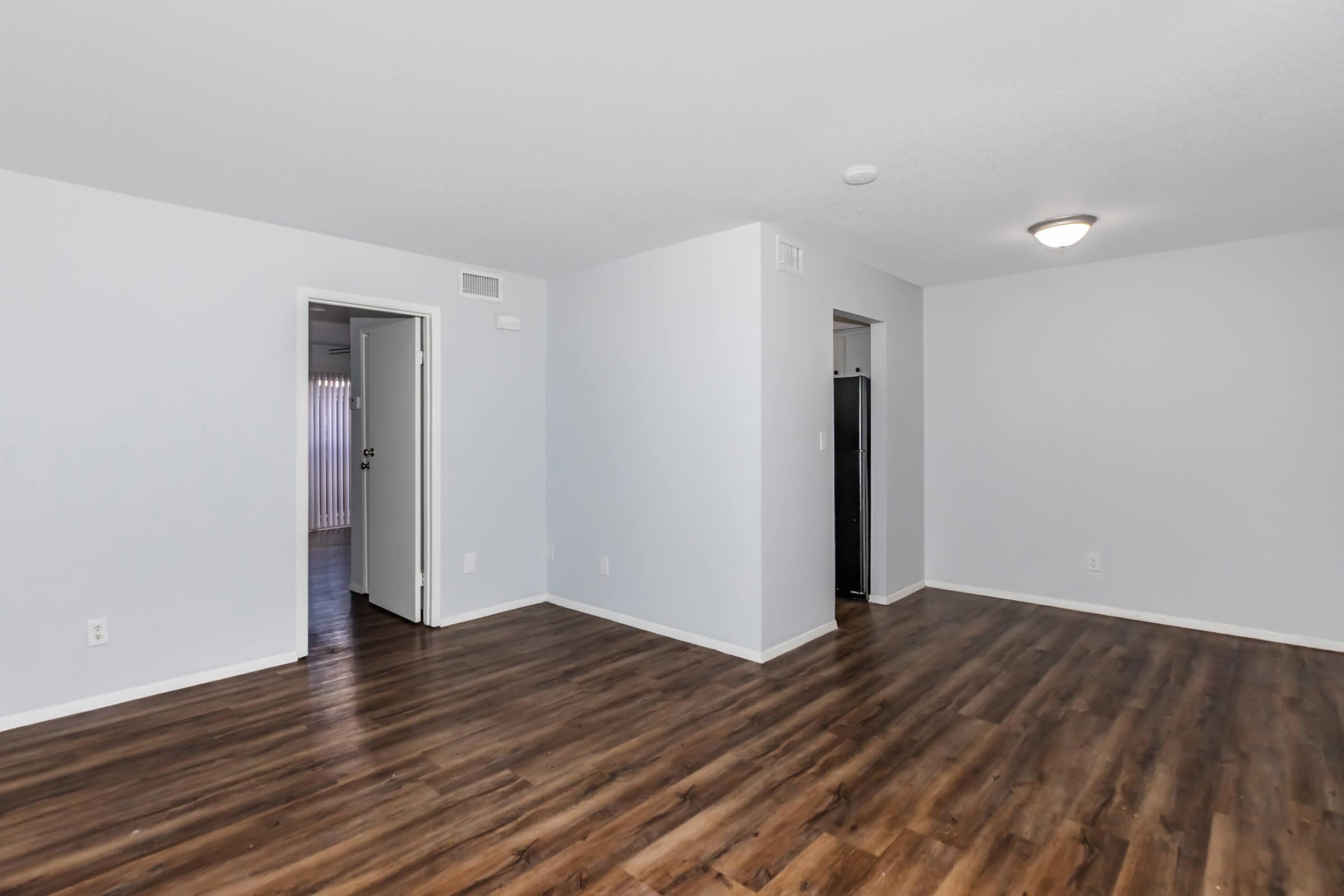 Spacious interior of an apartment featuring light gray walls, a wood-like floor, and multiple doorways. One doorway leads to another room, while a second door appears to lead to a kitchen or utility area. Natural light may be present, enhancing the welcoming atmosphere of the space.