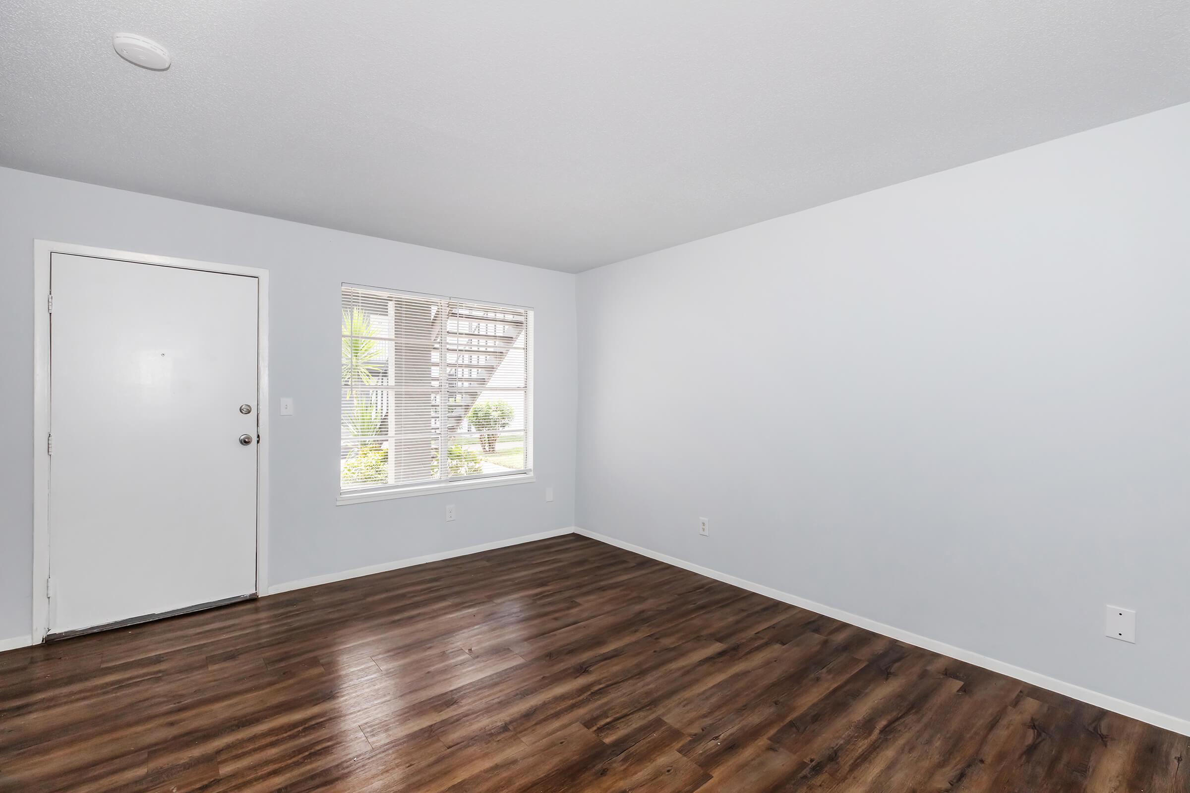 Interior of a room with light gray walls and a wood laminate floor. There is a white door on the left and a window with blinds on the right, allowing natural light to enter. The space is empty, creating a clean and minimalist look.