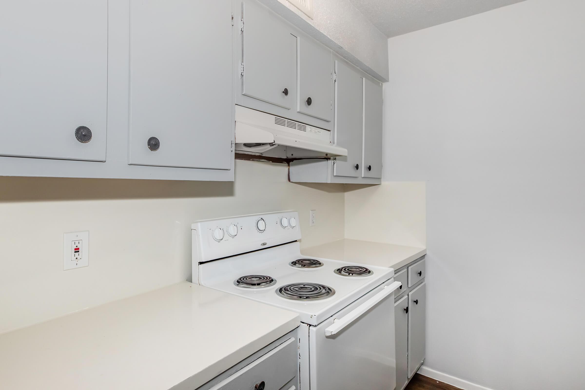 A modern kitchen featuring gray cabinets, a white electric stove with four burners, and a white countertop. The kitchen has a clean, minimalist design with an overhead range hood and light-colored walls, creating a bright and open space.