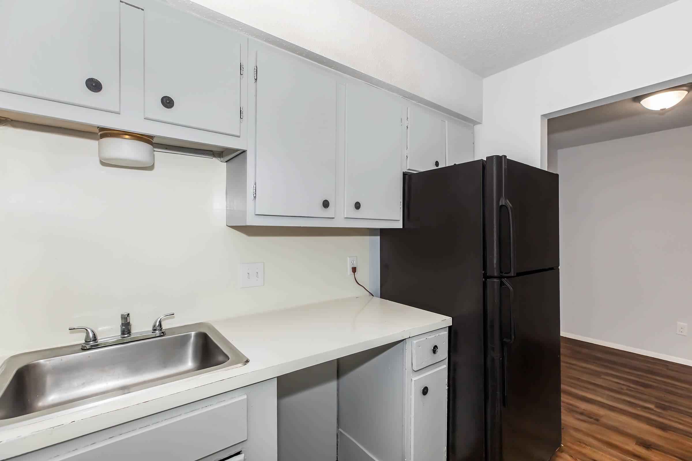 A kitchen view featuring gray cabinets, a stainless steel sink, and a black refrigerator. The countertops are white, and the floor displays wooden planks. The kitchen connects to a brightly lit room visible through an open doorway. The overall design is simple and modern.