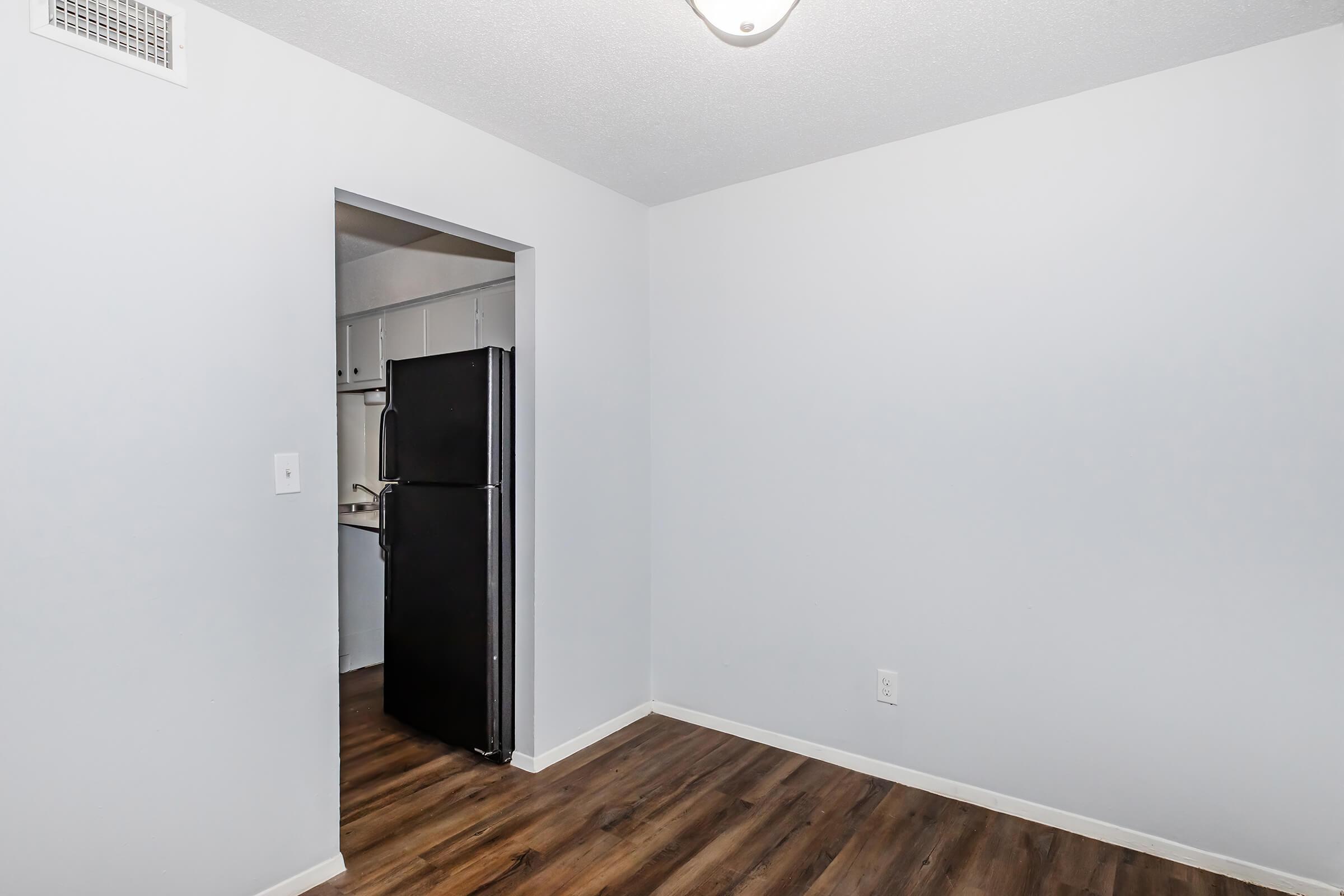 A view of a small room with light gray walls and dark wood laminate flooring. There is a doorway leading into another space, which appears to be a kitchen area with a black refrigerator visible. The ceiling has a light fixture. The room is empty and spacious.