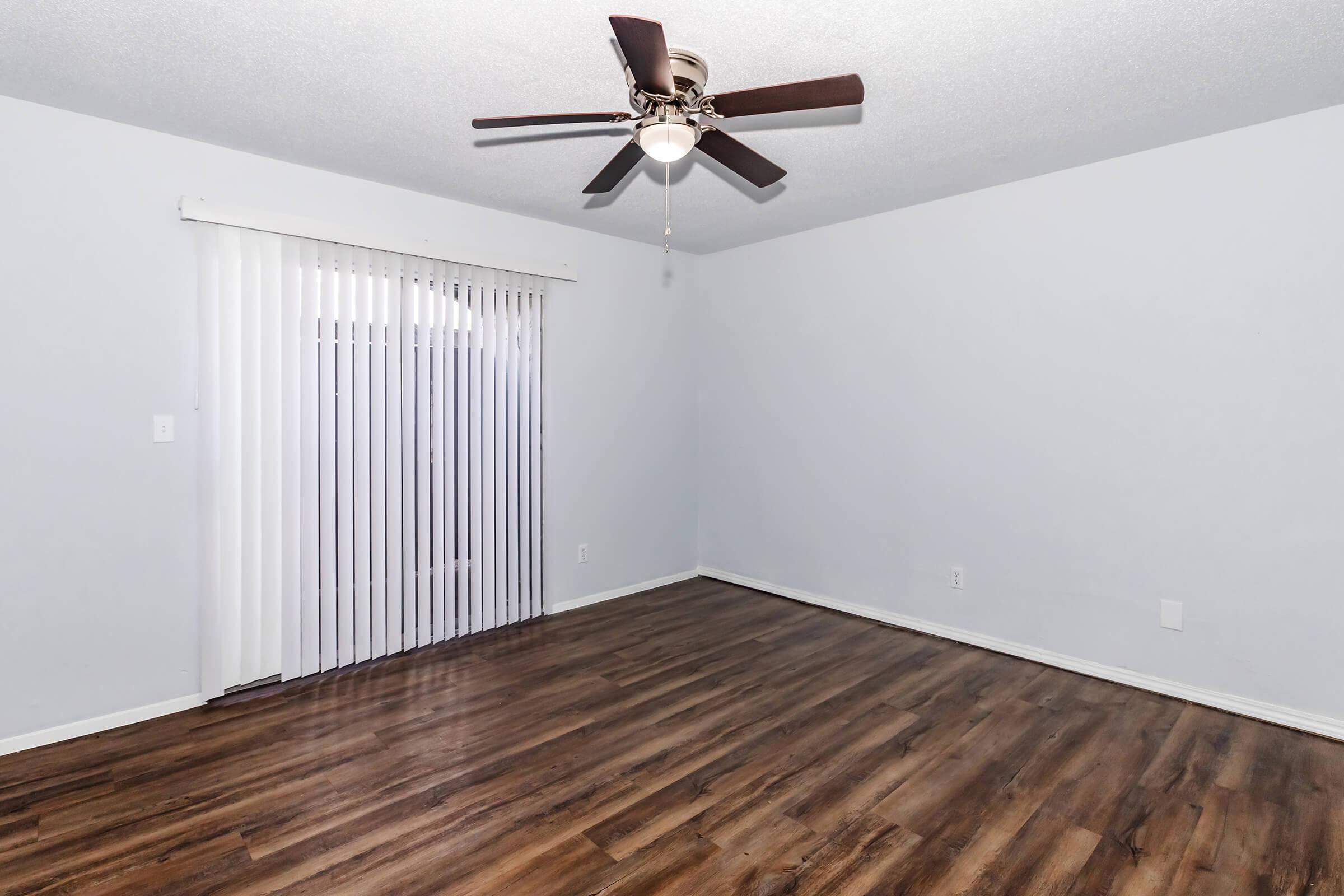 A spacious, empty room with light blue walls and a ceiling fan. The flooring is wooden, and there is a sliding glass door with vertical blinds on one side, leading to an outdoor area. The room has a bright and airy feel, suitable for various uses.