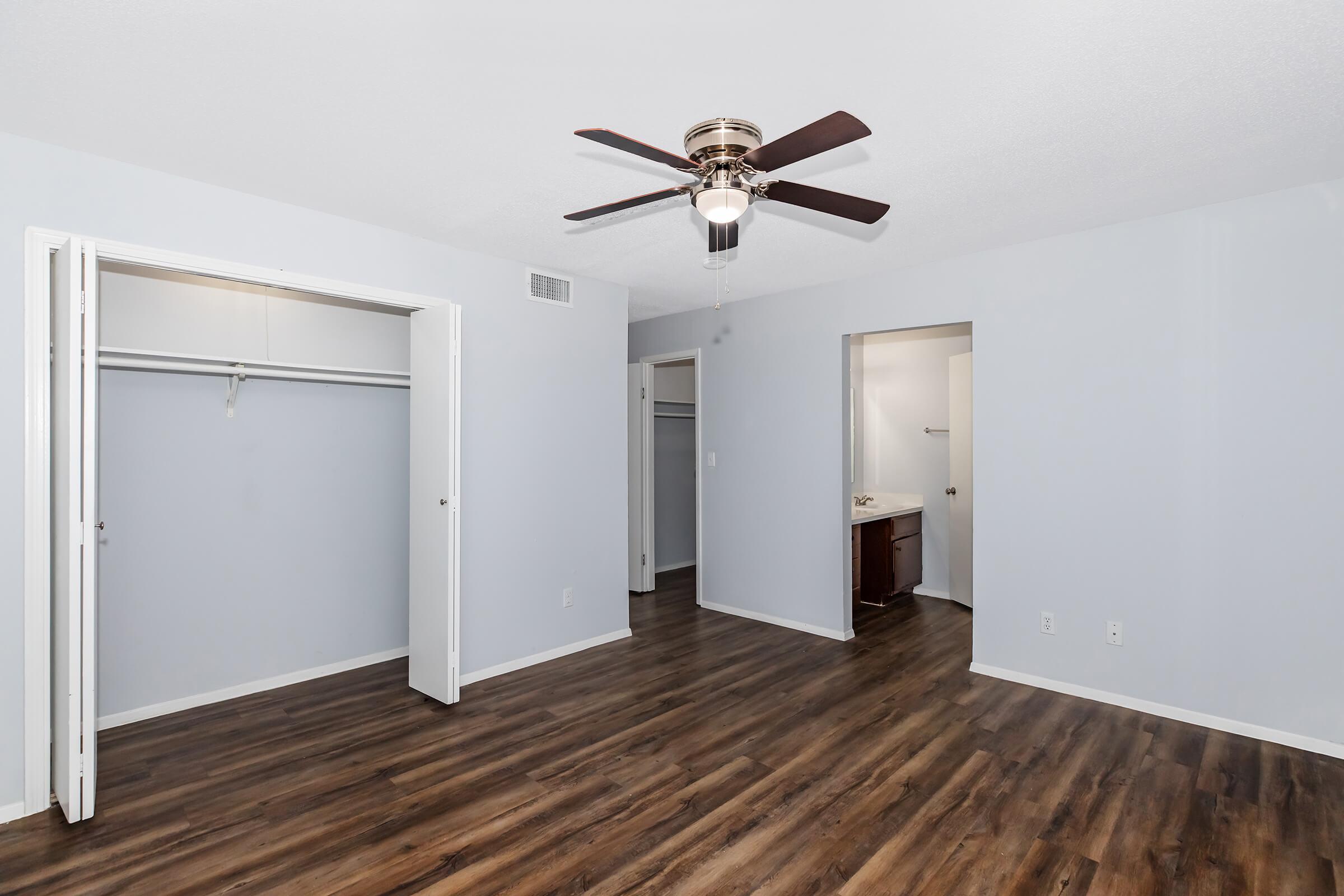 A spacious bedroom featuring light blue walls, wooden flooring, and a ceiling fan. Two doors lead to closets on the left, while an open doorway on the right indicates a bathroom. Natural light illuminates the room, creating a bright and airy atmosphere.