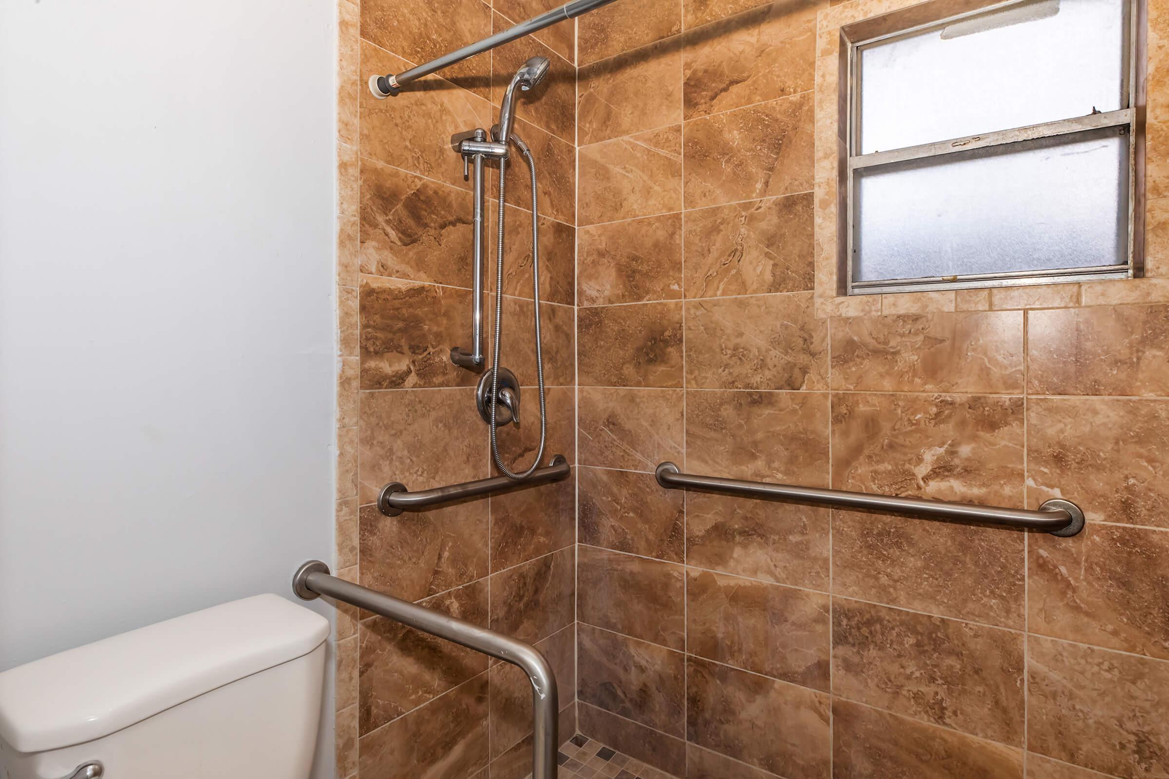 A modern bathroom featuring a walk-in shower with a grab bar, a wall-mounted showerhead, and a small window. The walls are adorned with large, textured brown tiles. To the left, there is a white toilet, and safety grab bars are installed for accessibility.