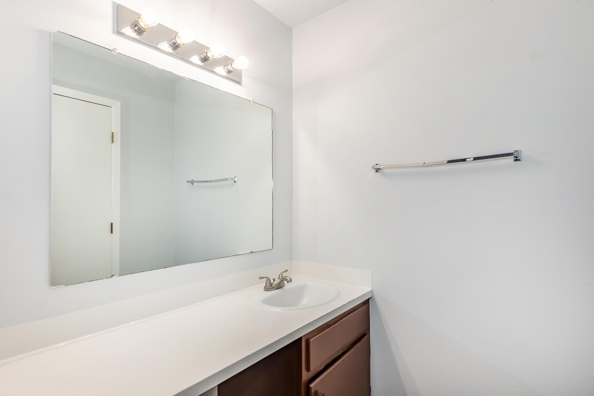A clean bathroom with a single sink and a large mirror above it, featuring four light fixtures mounted on the wall. The countertop is white, and there are wooden cabinets below. A towel bar is mounted on the wall next to the mirror, and the walls are painted white, creating a bright and simple space.