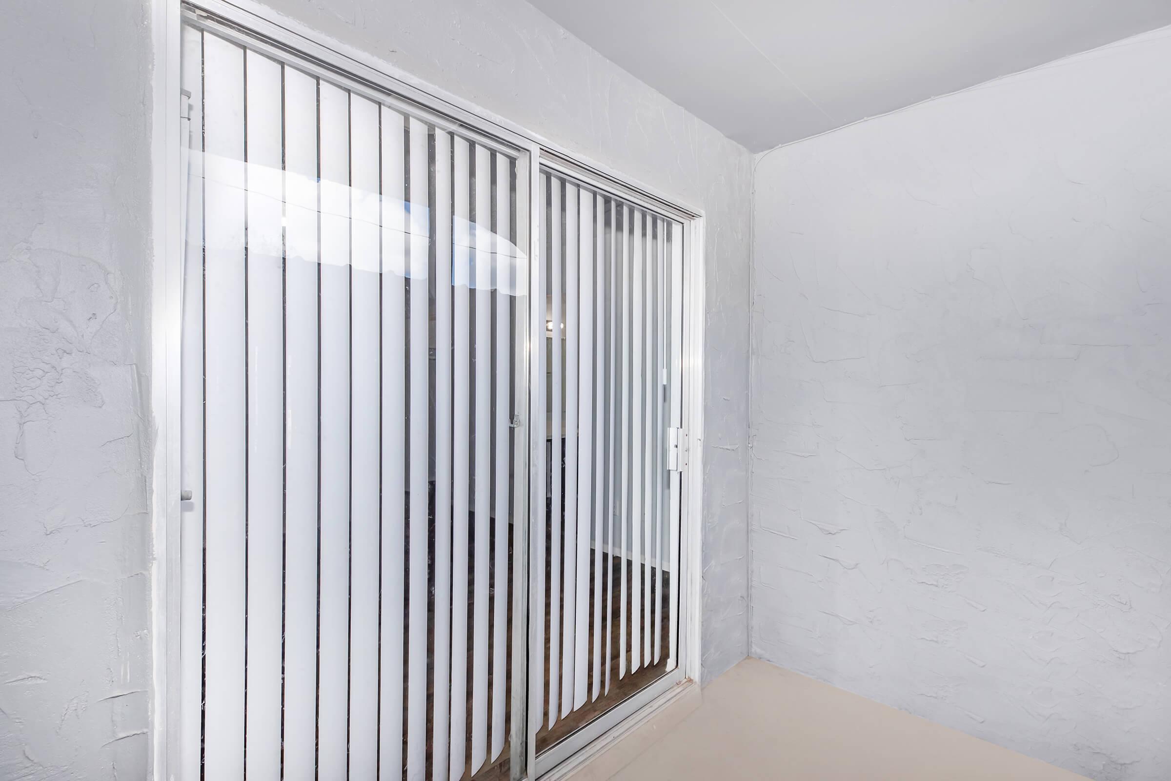 A corner view of a room featuring sliding glass doors with vertical blinds. The walls are painted in a light color, and the floor appears to be neutral-toned. Natural light is coming through the doors, illuminating the space.
