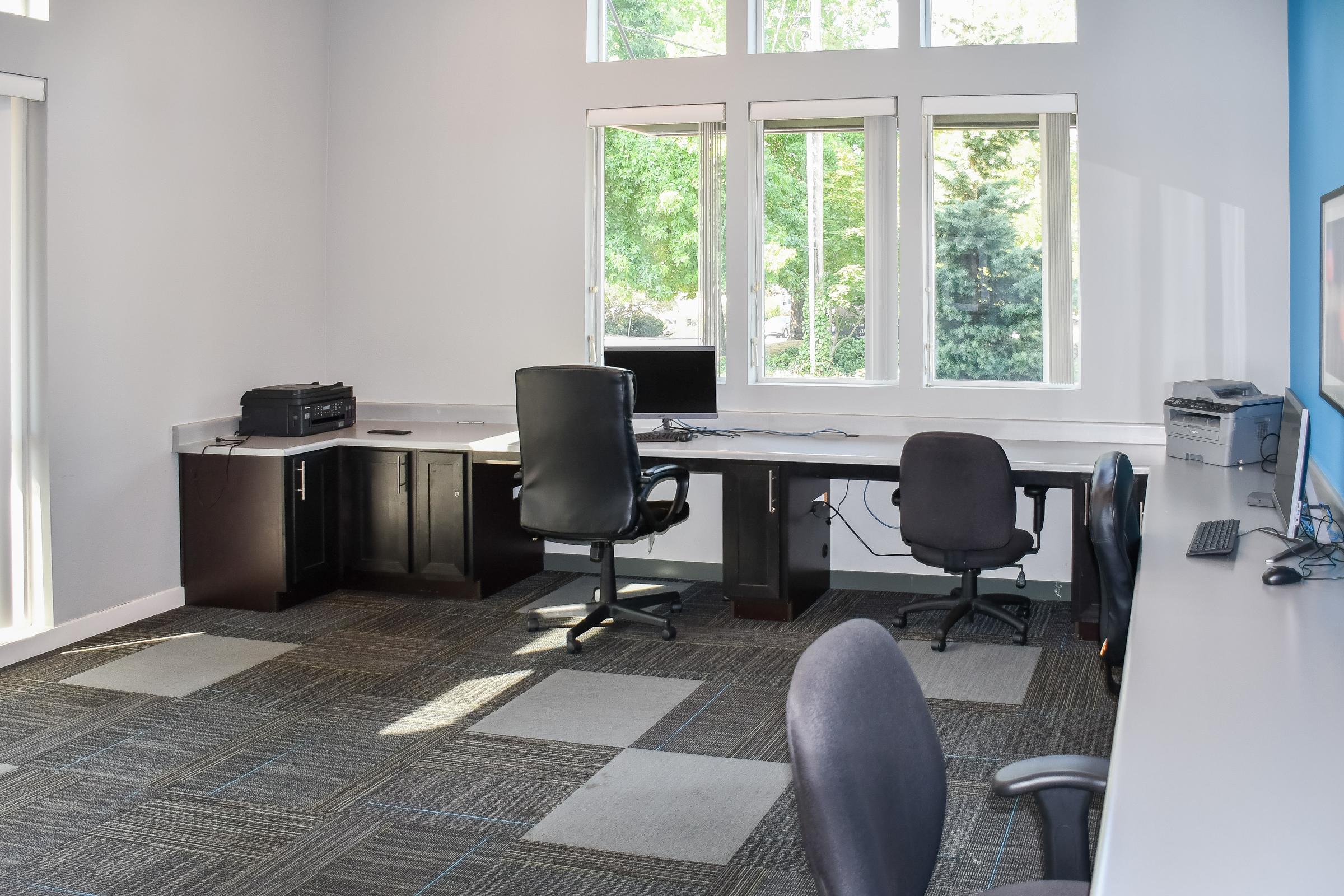 A bright, spacious office space featuring a large desk with two office chairs. There are large windows allowing natural light, and the room includes a printer and a computer setup on the desk. The floor is covered with carpet tiles in varying shades of gray.