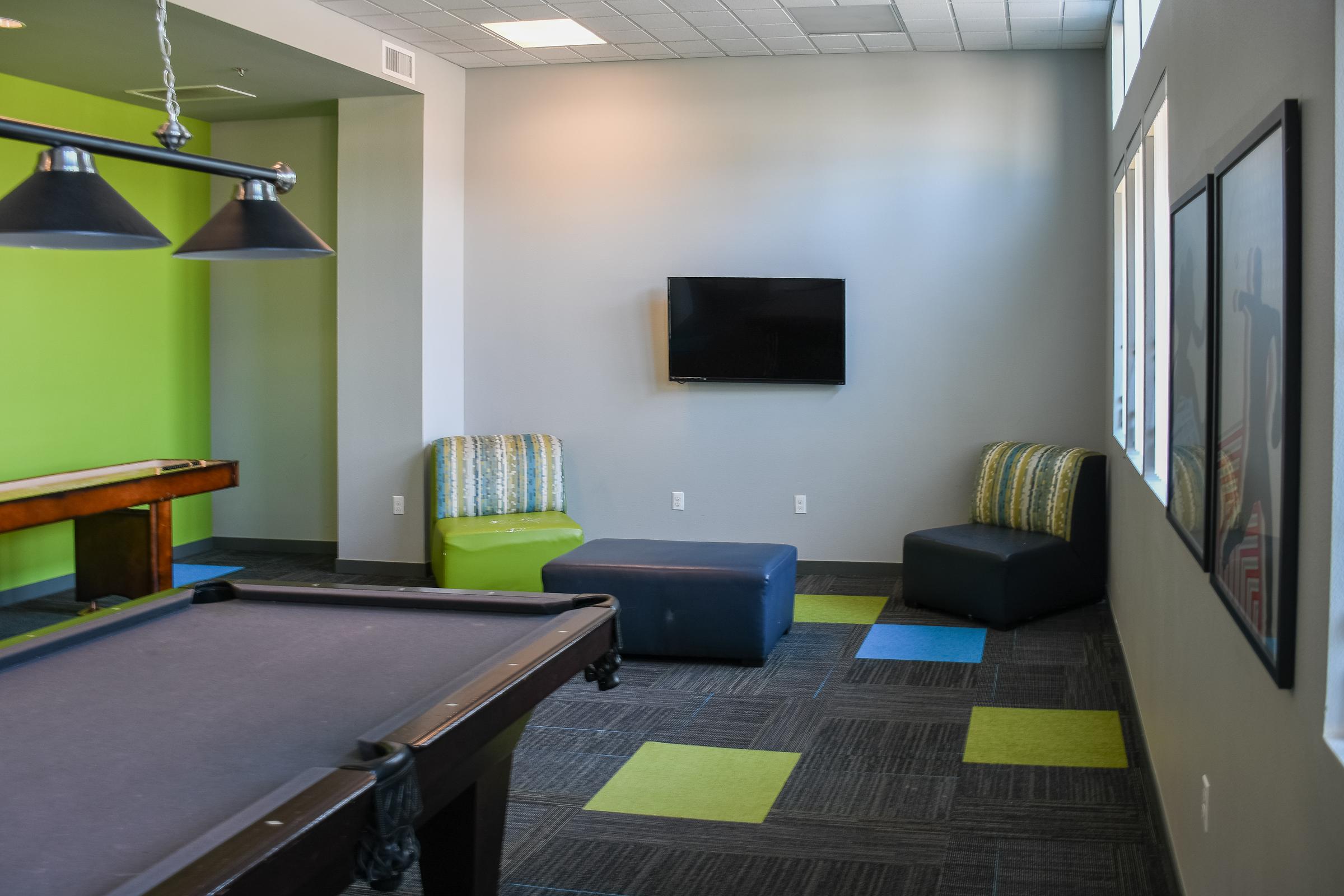 A recreational room featuring a dark billiard table, two stylish chairs in green and blue stripes, a dark blue ottoman, and a mounted flat-screen television. The walls are a mix of light gray and a vibrant green, complemented by a patterned carpet with colorful squares. Large windows let in natural light.