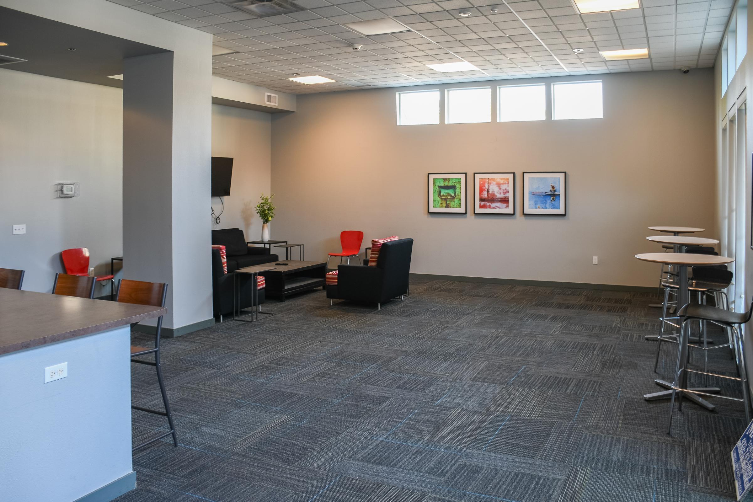 A spacious, modern lounge area featuring a dark couch, two red chairs, and a small table. Three framed artworks are displayed on the gray wall. The floor is covered with gray carpeting, and there are bar-height tables with stools on one side. Large windows provide natural light, creating an inviting atmosphere.