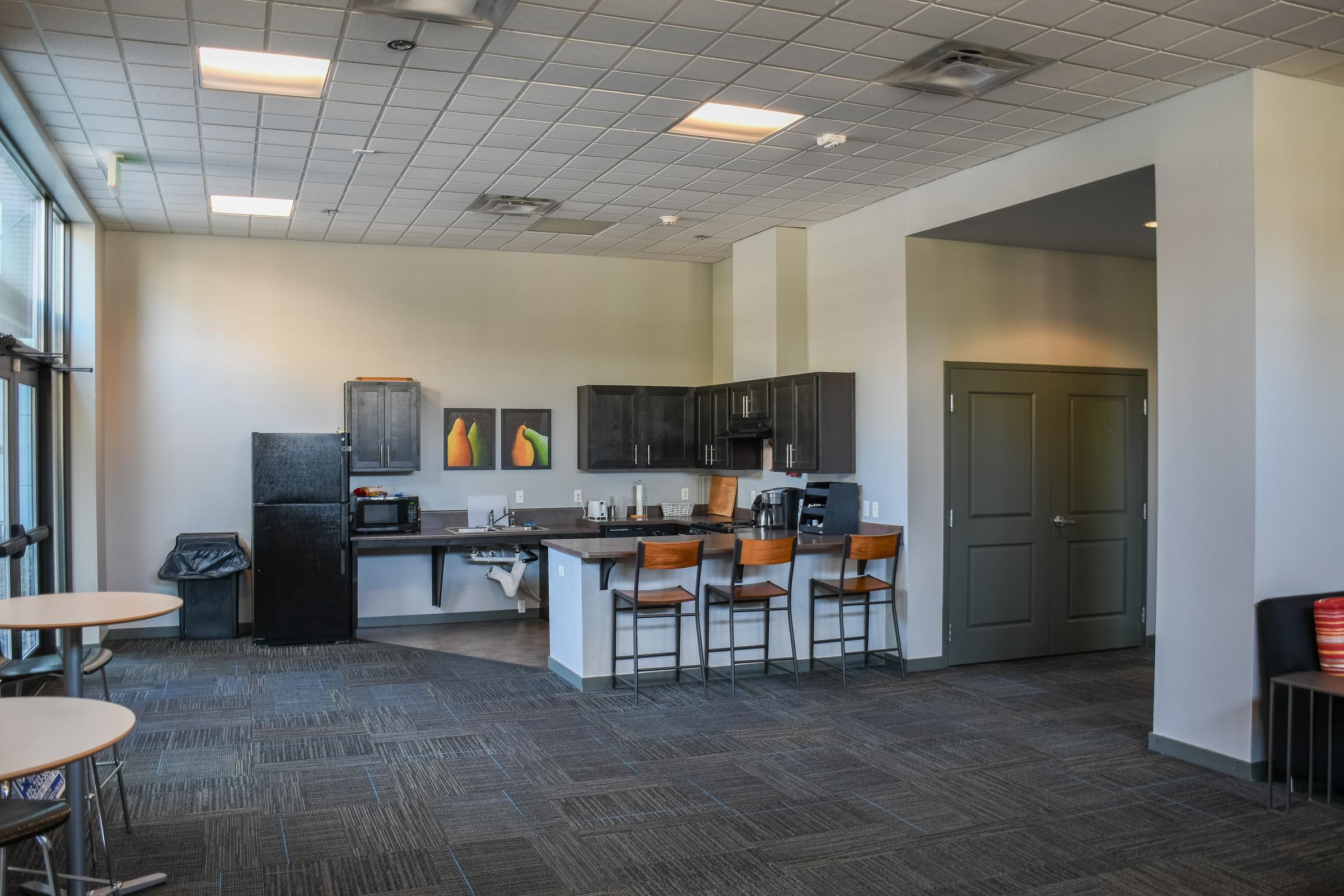 A modern kitchen area in a shared space featuring dark cabinetry, a countertop with bar stools, appliances including a microwave and oven, and artwork on the wall. The room has large windows allowing natural light, and a casual seating area with small tables. The flooring is carpeted with a textured pattern.