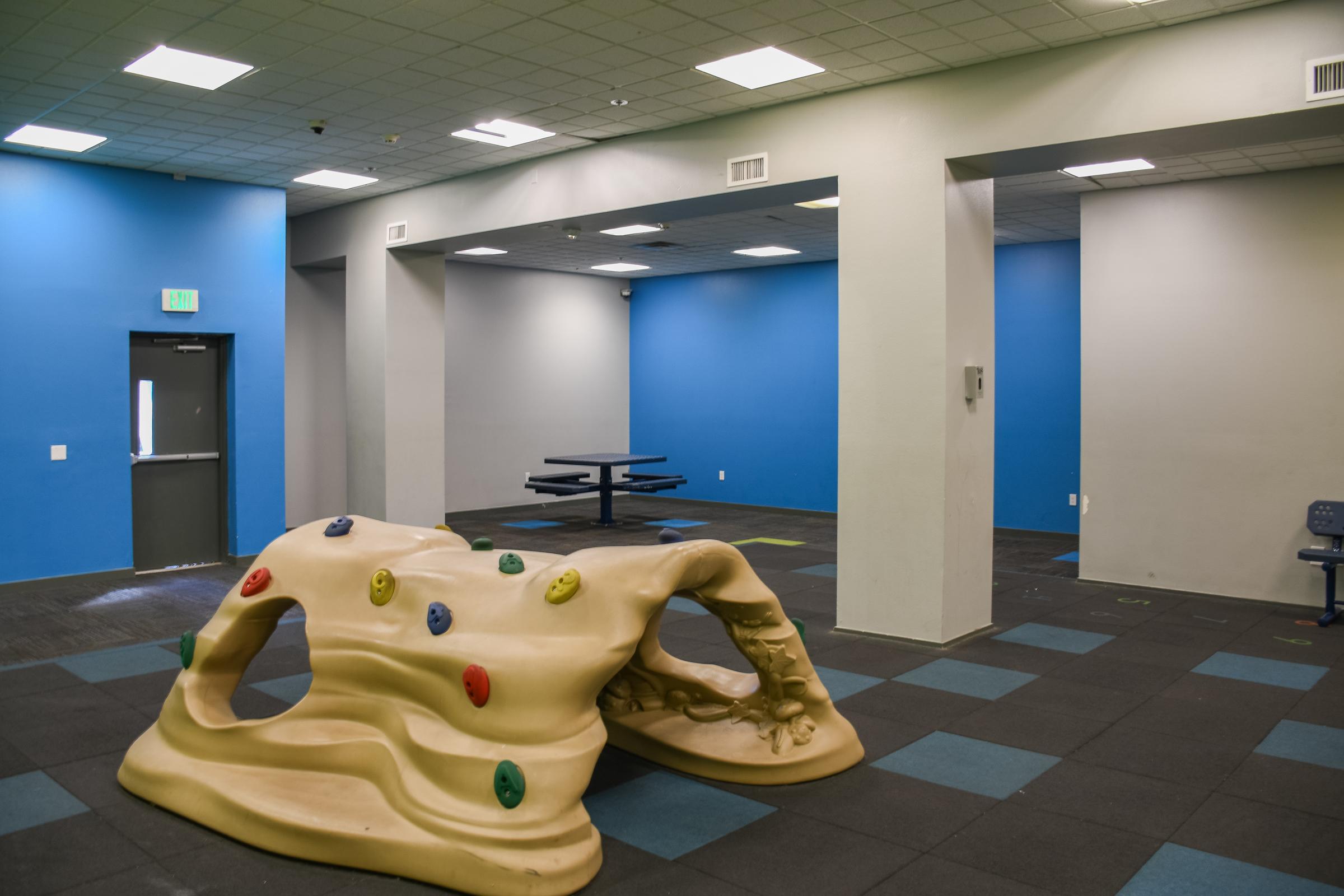 Indoor play area featuring a climbing structure with brightly colored handholds, set against blue-painted walls. In the background, there are tables and chairs, with a spacious layout on dark rubber flooring. The environment is designed for recreational activities.