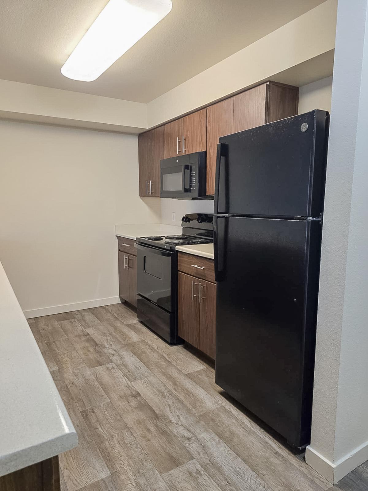 Modern kitchen featuring dark wood cabinets, a black refrigerator, and a black stove with a microwave above. Countertops are light-colored, and the flooring is a wood-like laminate. The space is well-lit with overhead lighting, and the walls are painted in a neutral shade.