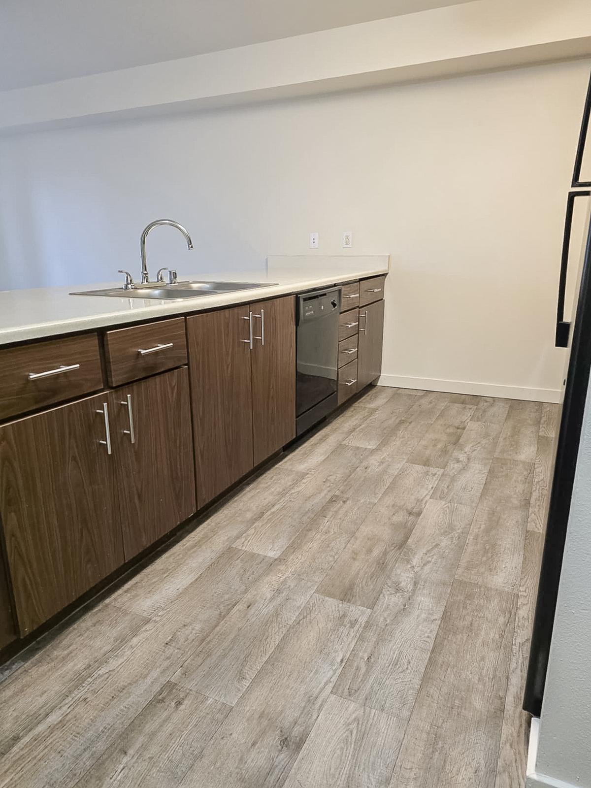 A modern kitchen featuring wooden cabinetry, a stainless steel dishwasher, and a sink. The floor is covered with light-colored laminate or hardwood, creating a spacious and clean aesthetic. The walls are painted in a light color, enhancing the bright and airy feel of the space.