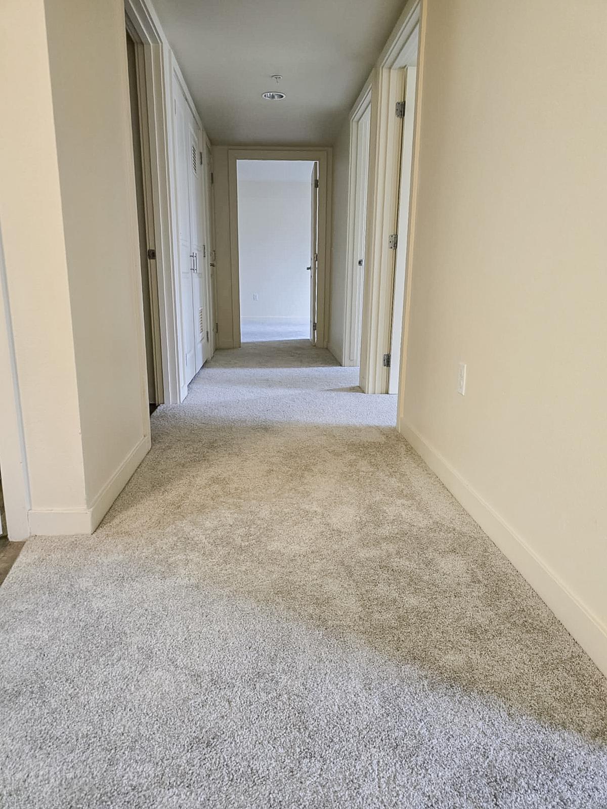 A view of a hallway with light-colored walls and a carpeted floor. The hallway extends into the distance, featuring several doorways on the left side and a visible door at the end. The lighting is bright and creates an open, spacious atmosphere.
