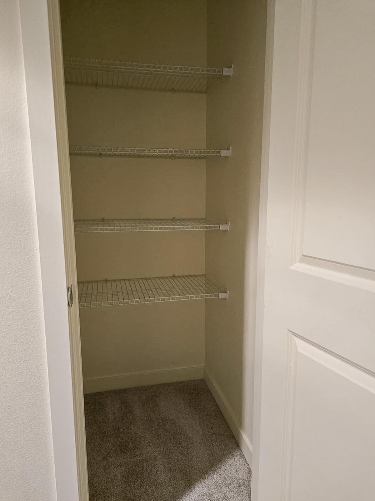 An empty closet with white wire shelving on four levels. The walls are painted a light color, and the floor is carpeted in a neutral tone. A white door is partially visible, showing the entrance to the closet space.