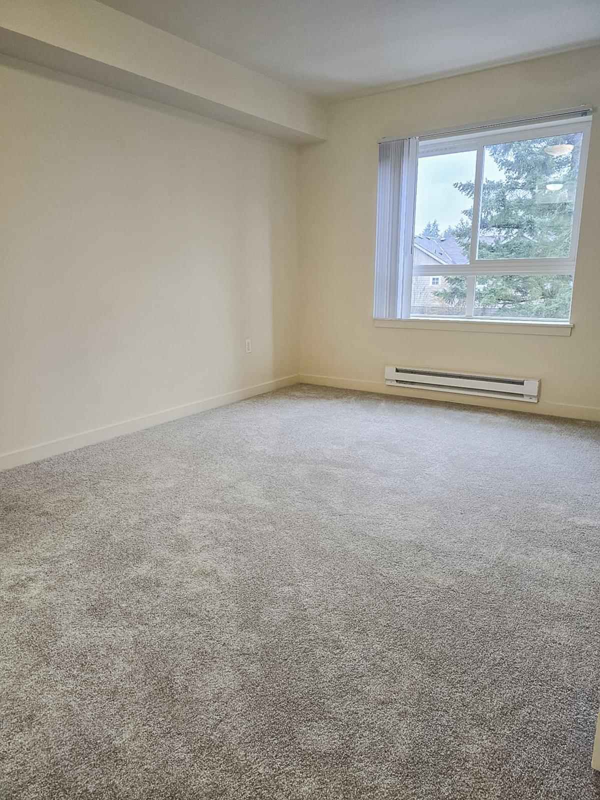 A spacious, empty room with light gray carpet and white walls. A large window with vertical blinds allows natural light to enter, highlighting the clean, open layout. The room features a heating unit along the baseboard and is devoid of furniture or decorations.