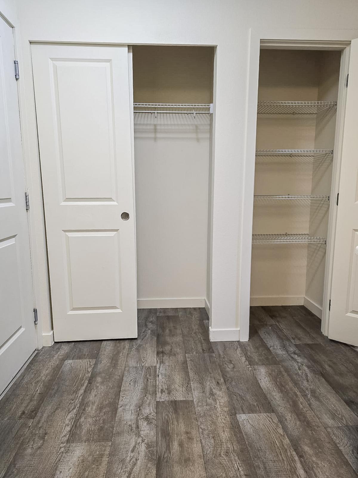 A well-lit, empty room featuring two open closet doors. One closet has a wire shelf and is partially visible, while the other closet is fully open with no items inside. The flooring is a modern, dark wood style, contributing to a clean and spacious atmosphere.