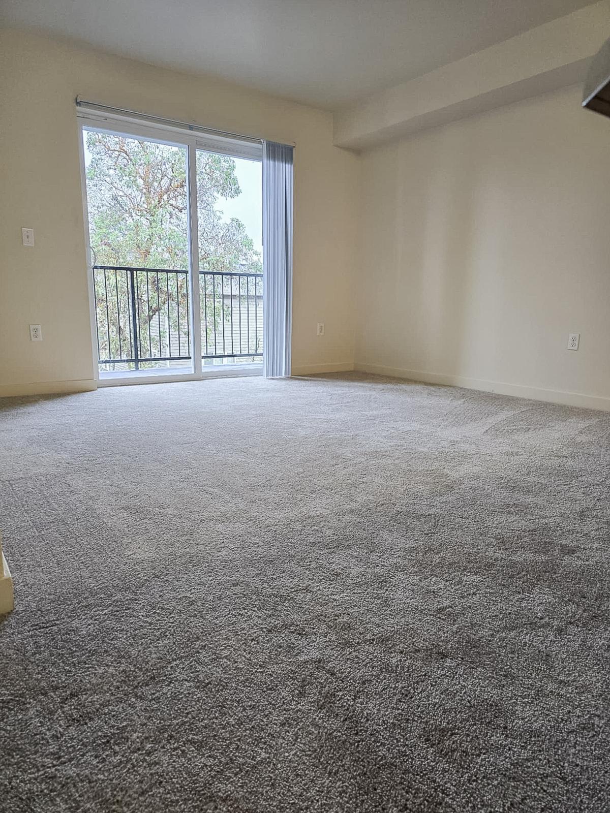 A spacious, empty room with light beige walls and gray carpet. A sliding glass door leads to a balcony, visible in the background, showcasing greenery outside. The overall atmosphere is bright and airy, with natural light flooding the space.