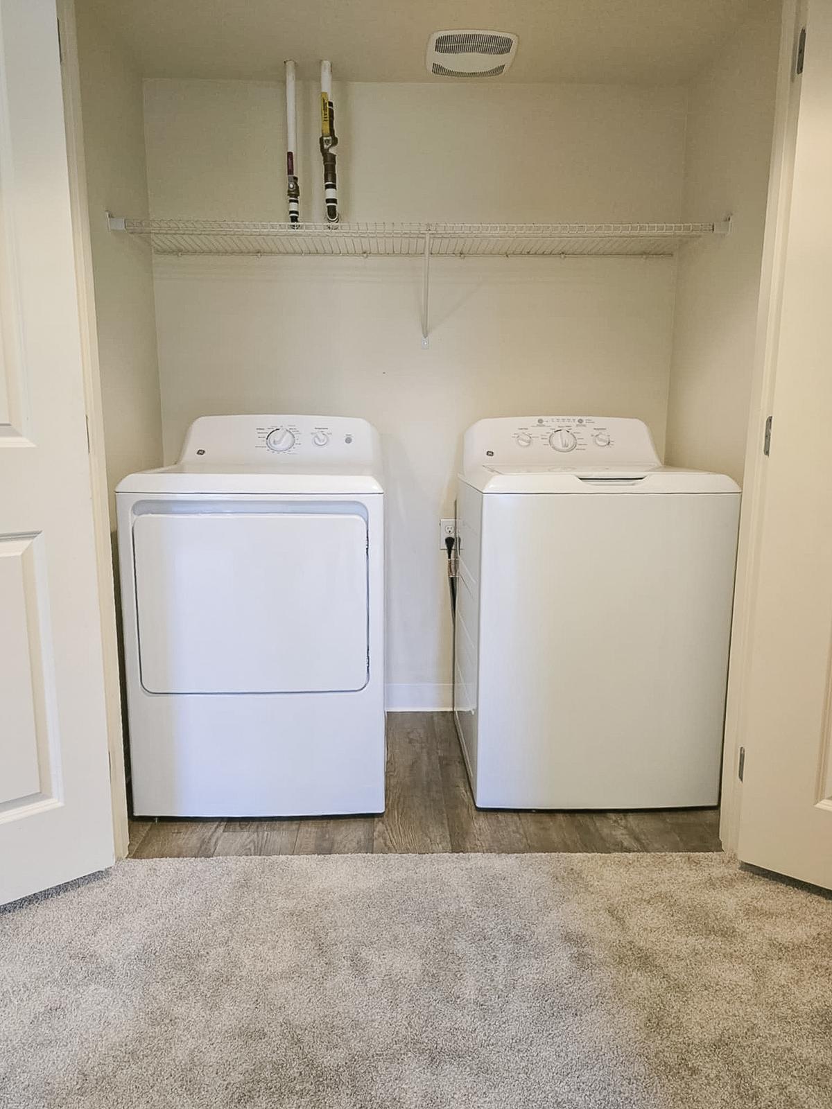 A laundry room featuring a stacked washer and dryer side by side, with a shelving unit above. The floor is carpeted, and the walls are a light color, creating a tidy and organized space.