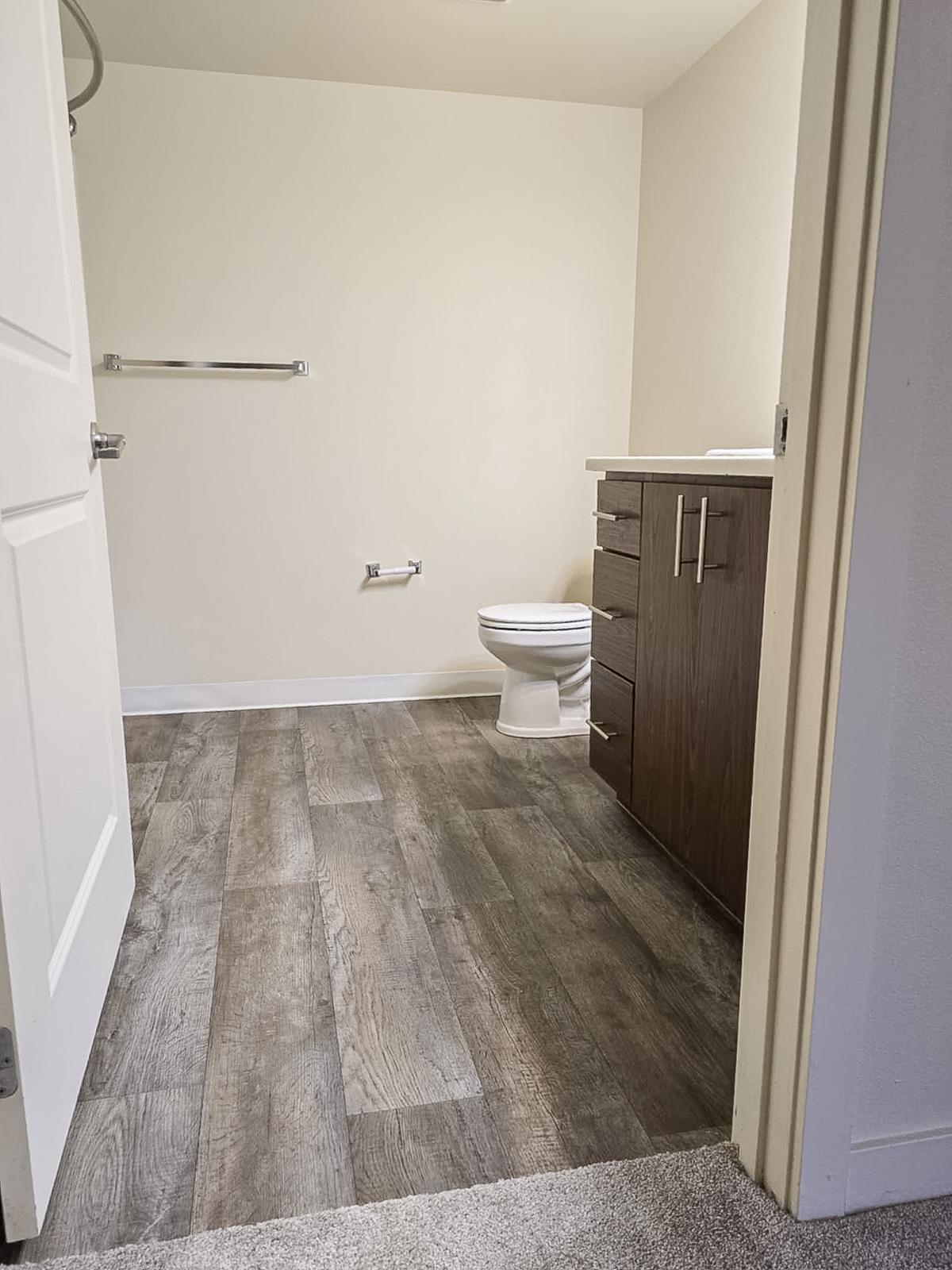 A clean, modern bathroom featuring a white toilet, a wooden vanity with drawers, and a towel bar. The floor is covered in wood-style laminate, and the walls are painted in a light color. The room is well-lit and minimalistic, with an open door providing access.