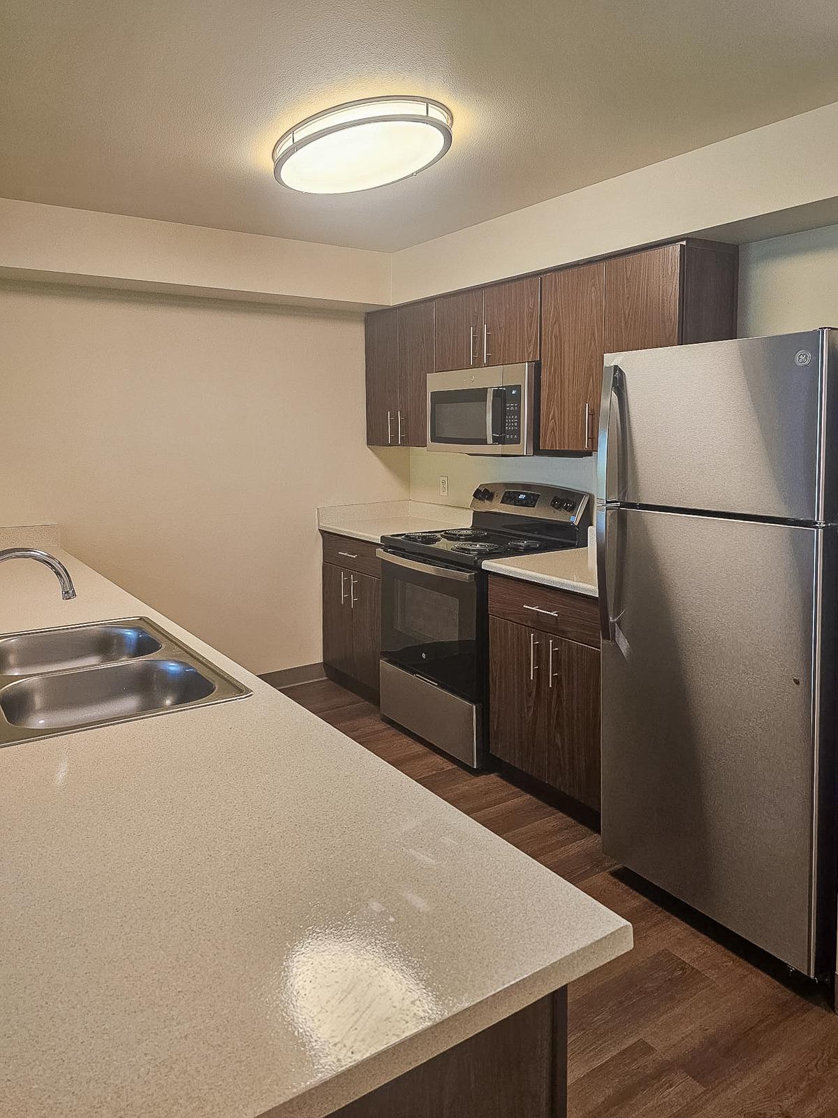 A modern kitchen featuring dark wooden cabinets, a stainless steel refrigerator, a microwave above the stove, and a double sink. The countertops are light-colored and smooth, with a sleek design. A round ceiling light fixture provides bright illumination in the space.