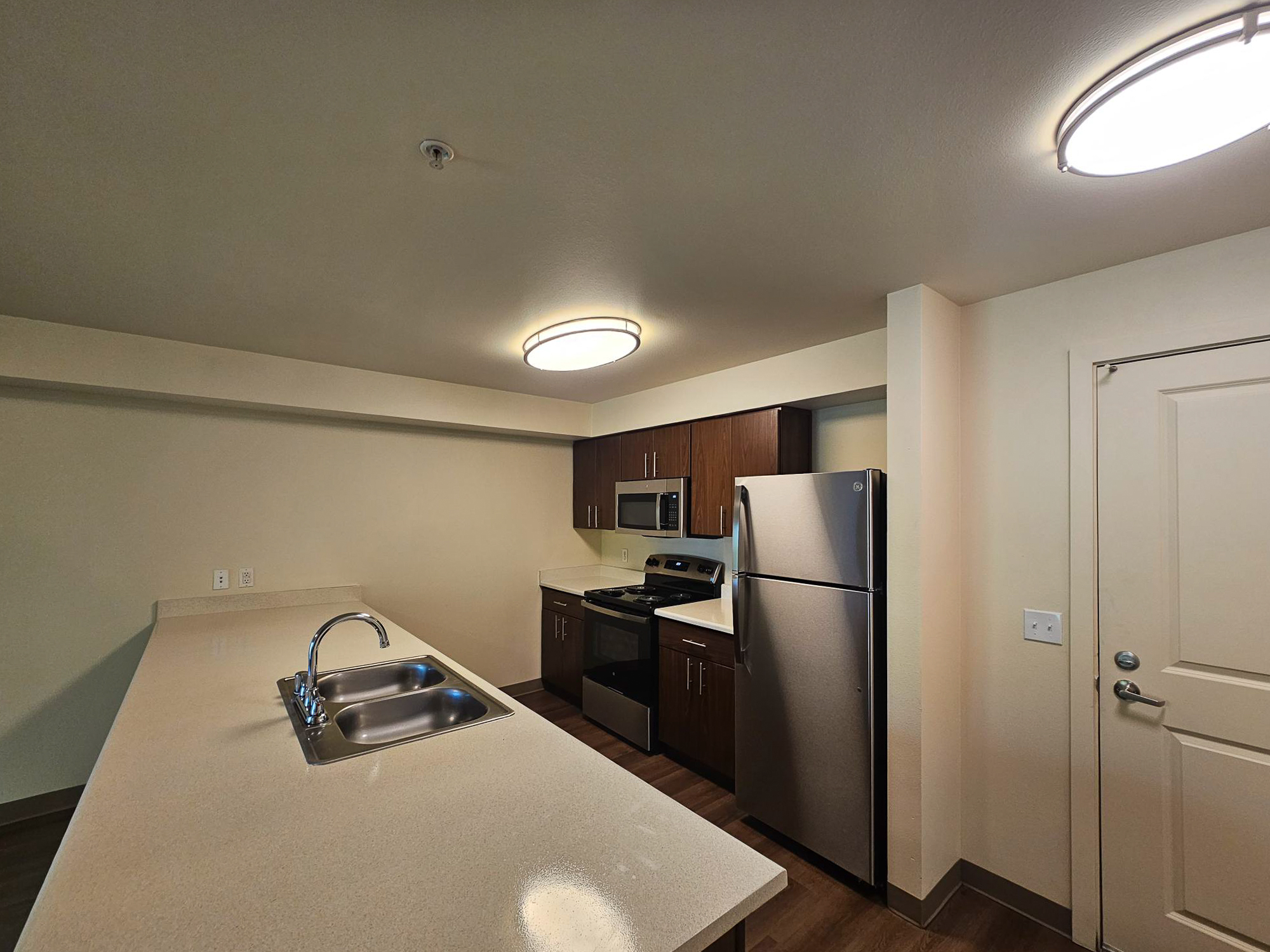 Modern kitchen featuring a center island with a double sink, stainless steel appliances including a refrigerator, oven, and microwave, dark wooden cabinets, and bright lighting from overhead fixtures. The walls are a soft neutral tone and there is a door leading to another room.