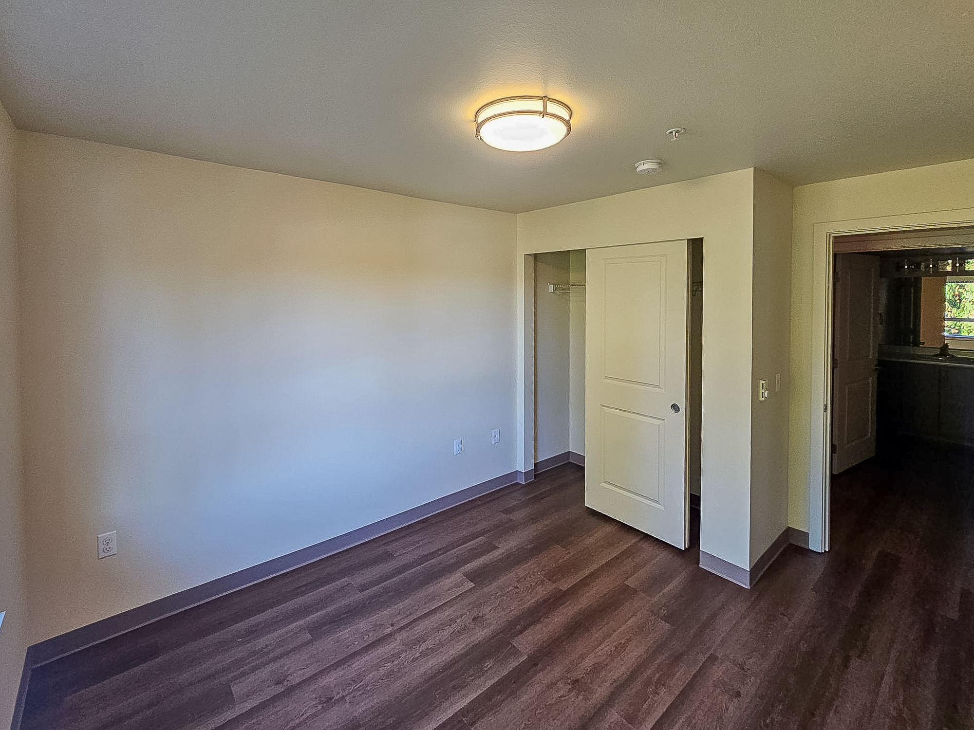 A spacious, empty room with light-colored walls and a modern ceiling light fixture. The floor is wooden, and there is a closet space with sliding doors on the left. A partially open door on the right leads to another room, suggesting an adjoining space. The overall atmosphere is bright and inviting.