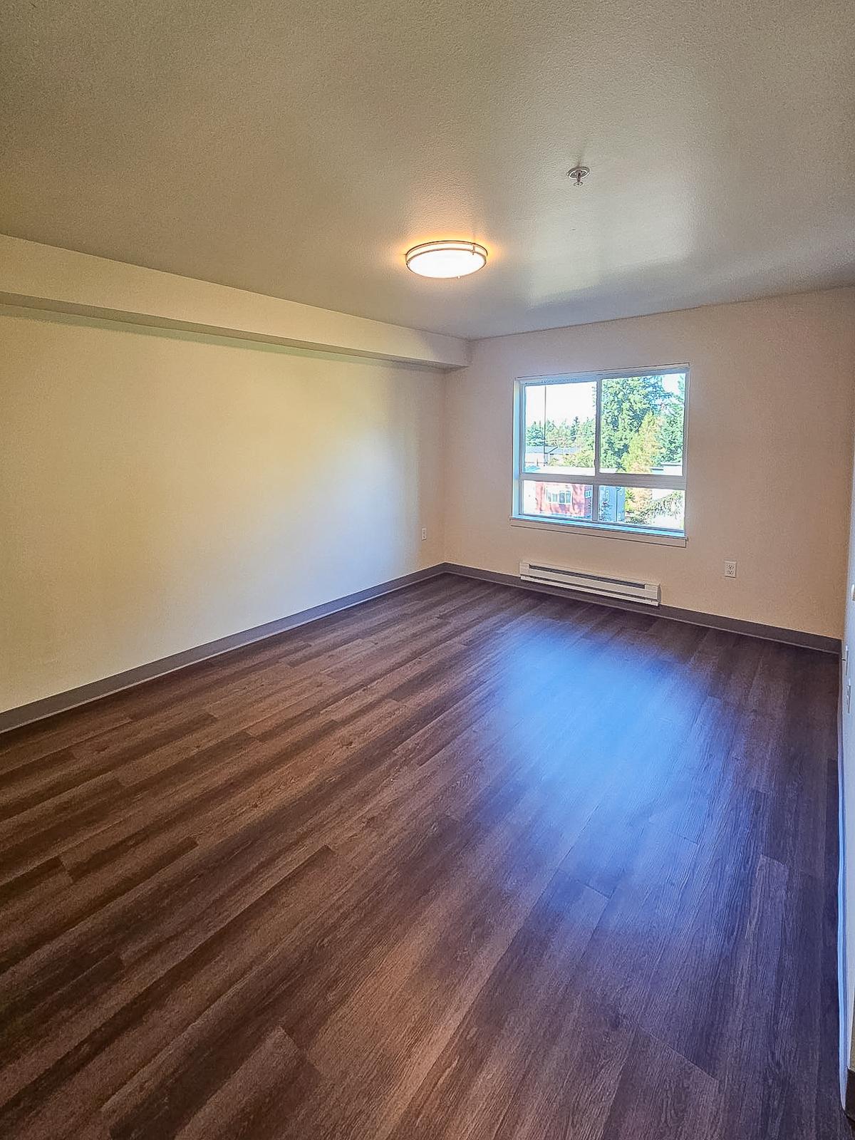 Empty room with light wood laminate flooring and a large window allowing natural light. The walls are painted in light colors, and there is a ceiling light fixture. The space is unfurnished, giving it a spacious and open feel.