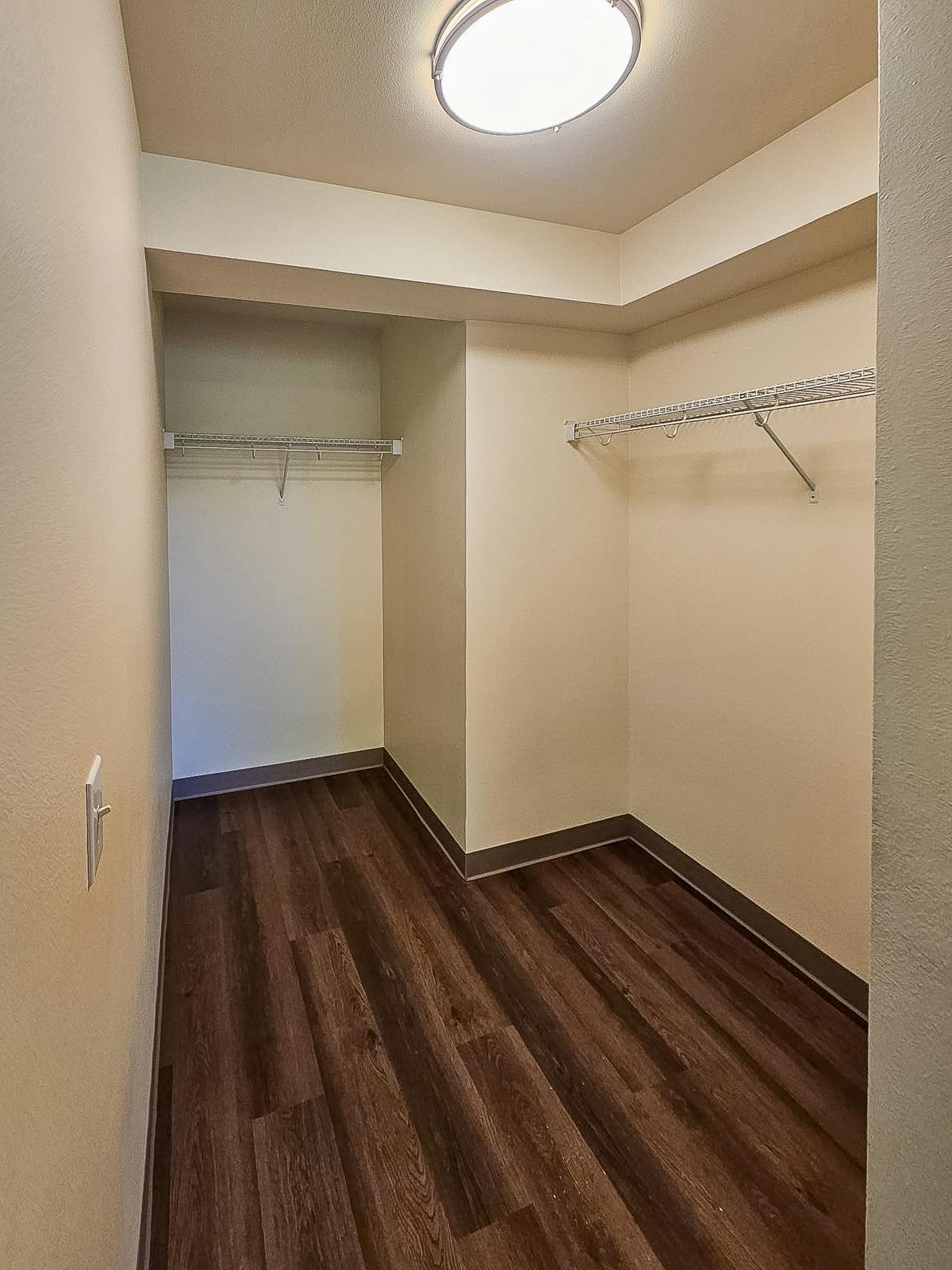A spacious, well-lit walk-in closet with beige walls, a ceiling light fixture, and dark wood-like flooring. Two wire shelves are mounted on the walls, providing storage options. The closet appears empty, creating a clean and organized space.