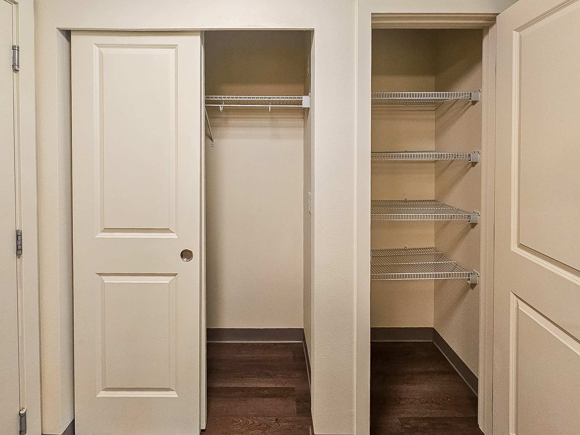 A well-lit hallway featuring two open closet doors. The left closet has a single hanging rod, while the right closet has multiple wire shelves. The walls are painted in a neutral color, and the floor is finished with dark wood.