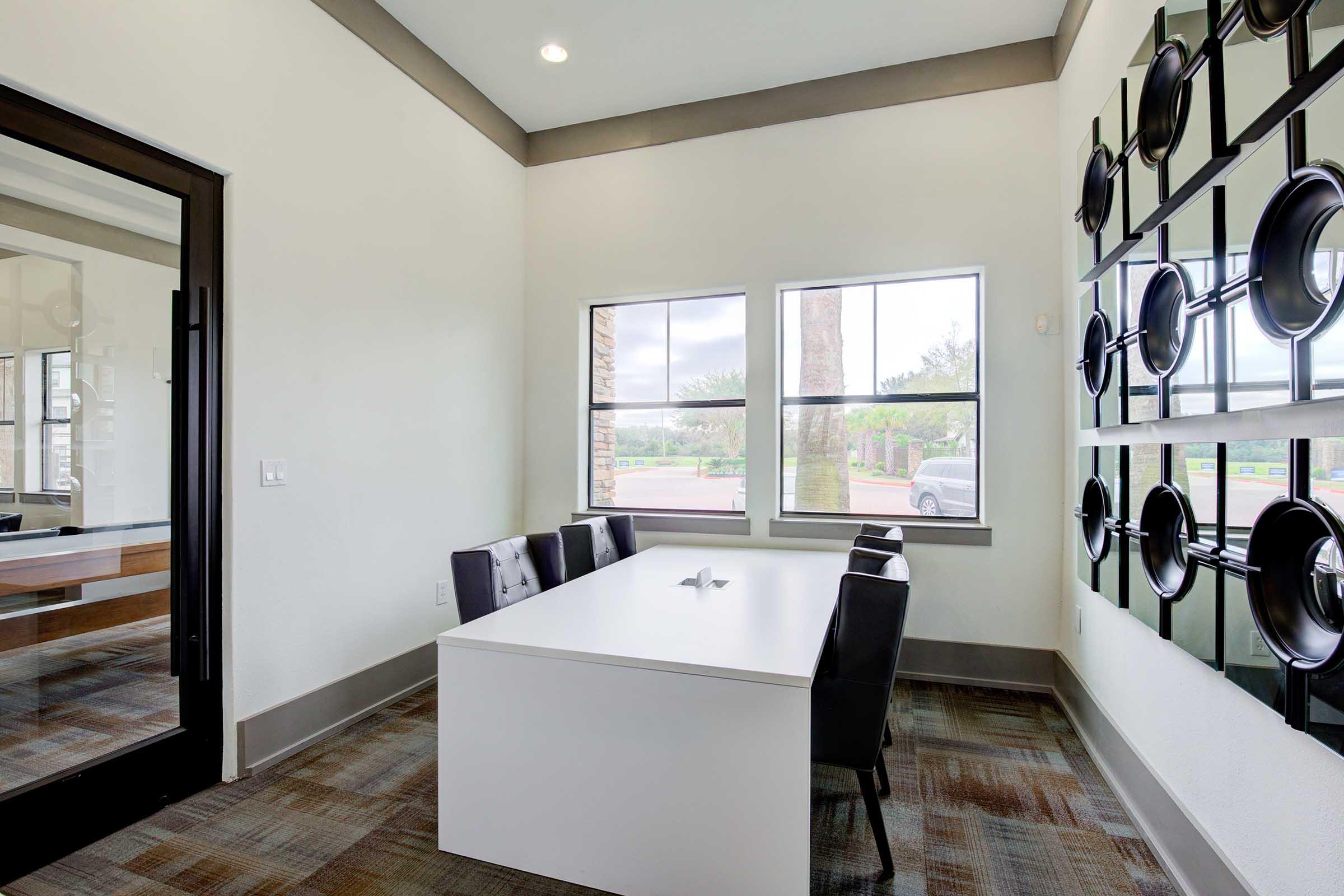 A modern, well-lit conference room featuring a white table surrounded by black chairs, with large windows showing trees outside. The walls are painted in light colors, and one wall has a decorative arrangement of circular black wall art. The space is uncluttered and inviting for meetings.