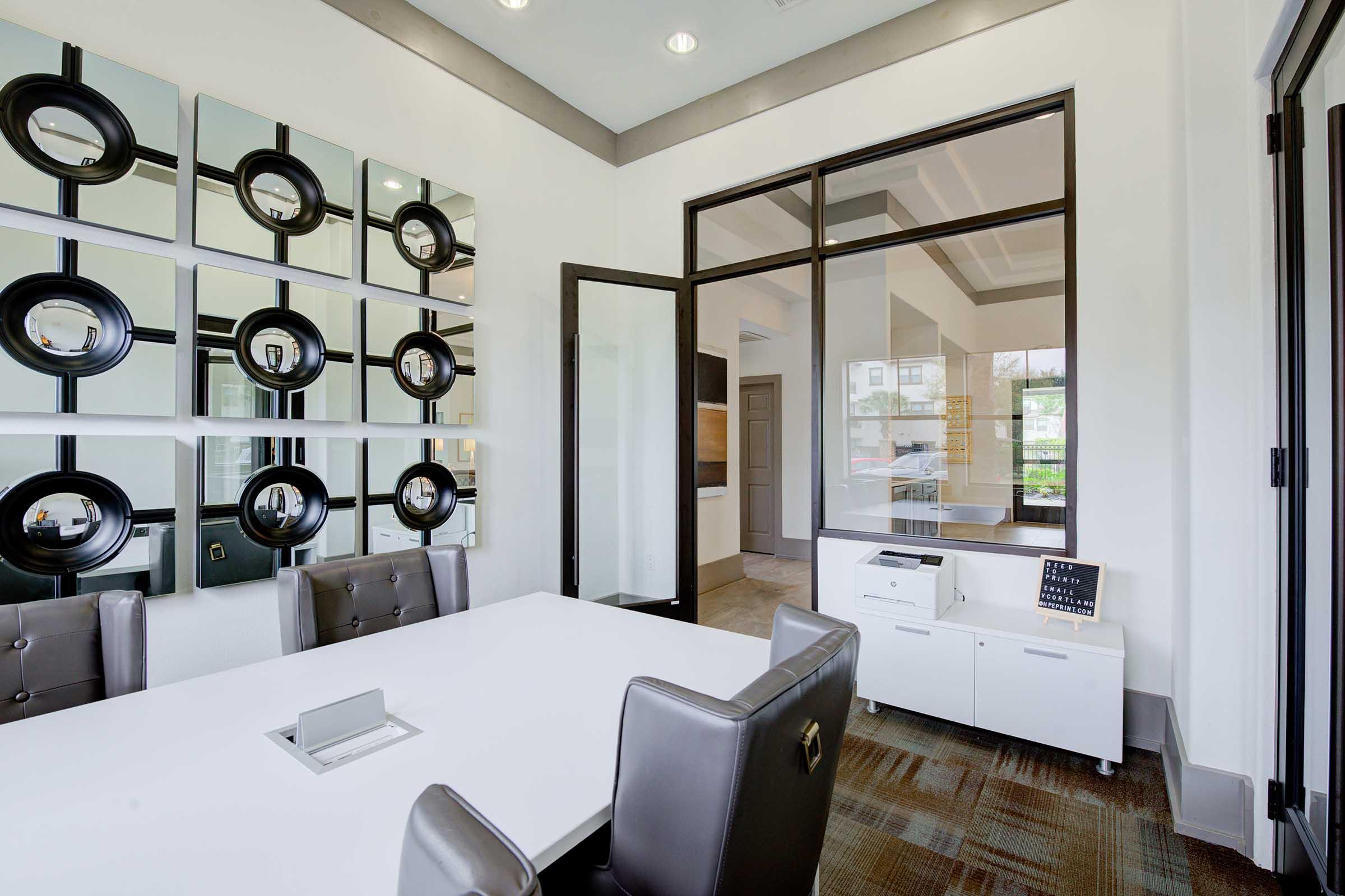 A modern office space featuring a sleek white conference table surrounded by gray chairs. On the walls, there are decorative mirrors arranged in a grid pattern. A large window provides natural light, and a printer is visible on a side cabinet. The overall design is contemporary and minimalistic.