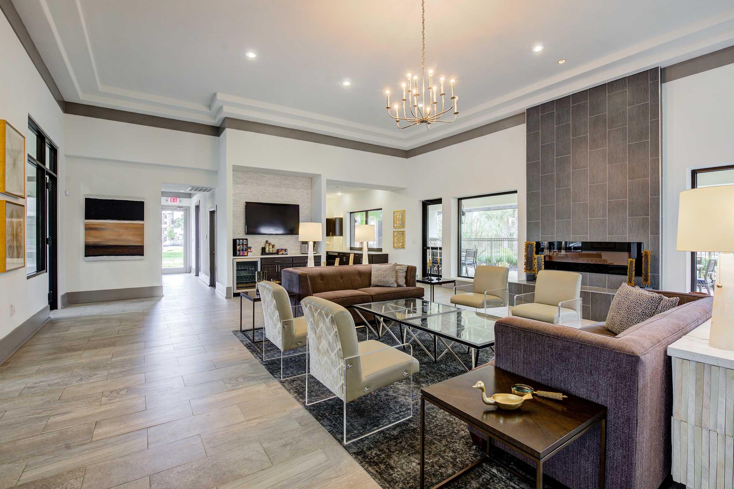 A modern living room featuring a stylish arrangement of sofas and chairs around a glass coffee table. The space is well-lit with a chandelier and large windows, showcasing a neutral color palette and contemporary decor, including artwork on the walls. There's a sleek fireplace and a television in the background.