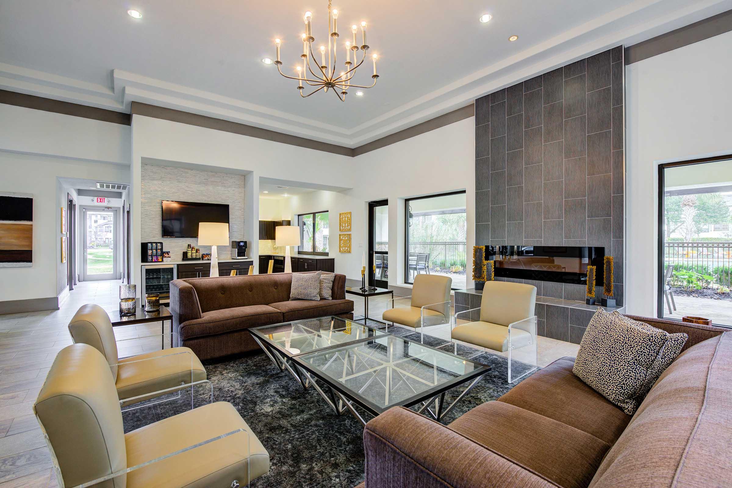 A modern living room featuring a large glass coffee table at the center, surrounded by various seating options including two brown sofas and clear armchairs. A stylish chandelier hangs from the ceiling, and a fireplace with a dark tiled backdrop adds warmth to the space. Large windows provide natural light.