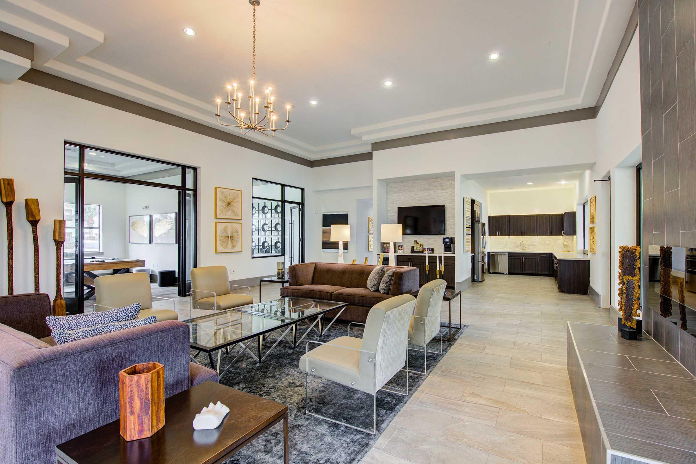 A spacious, modern living room featuring a plush seating area with a brown sofa and light-colored chairs. A coffee table sits on a dark area rug, and large windows allow natural light. The adjacent kitchen is visible, showcasing dark cabinets and light countertops, while decorative artwork adorns the walls.