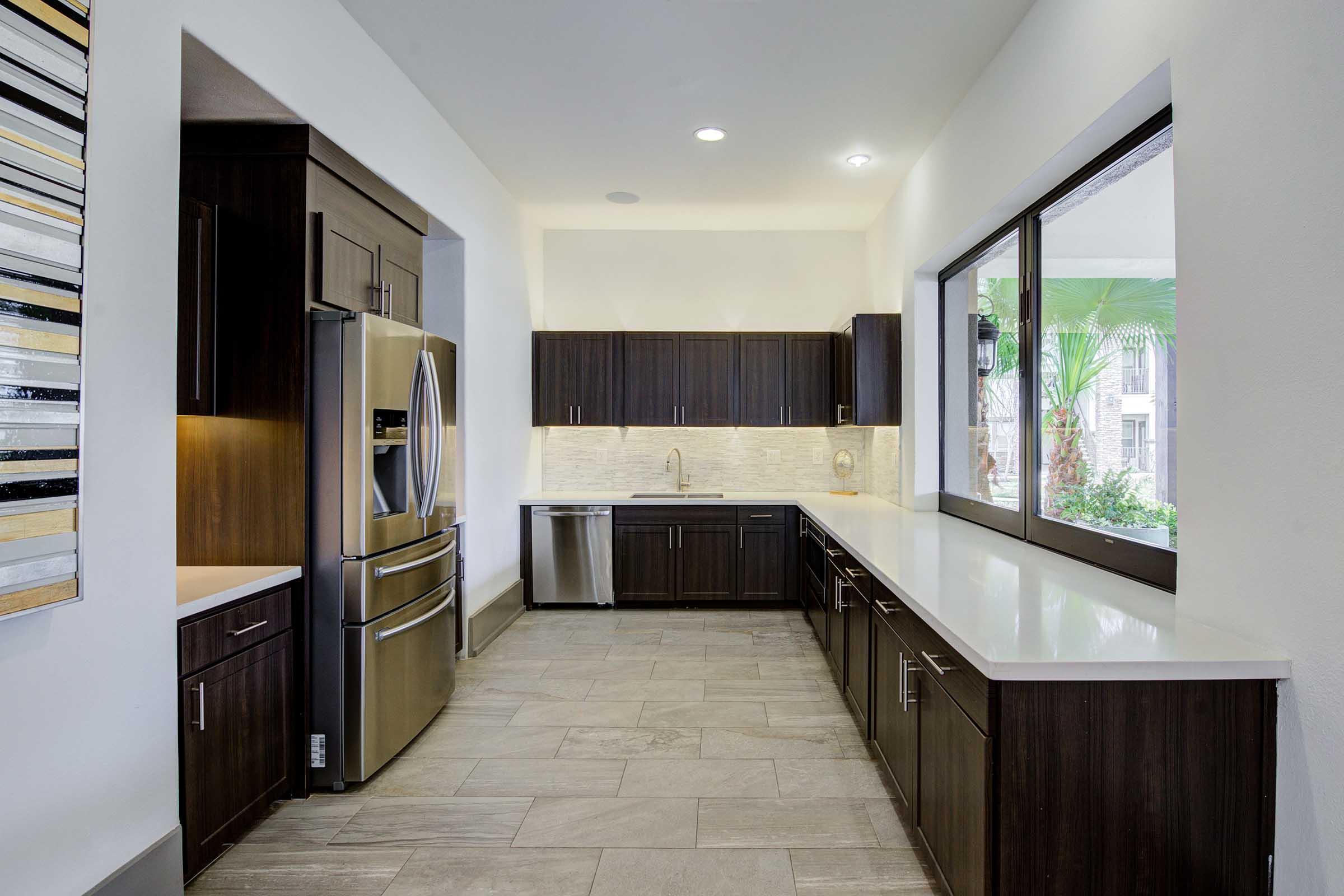 Modern kitchen featuring dark wood cabinets, a stainless steel refrigerator, and a sink with a window overlooking greenery. The space has light-colored countertops and tiled floors, creating a bright and inviting atmosphere. Natural light fills the area from large windows.