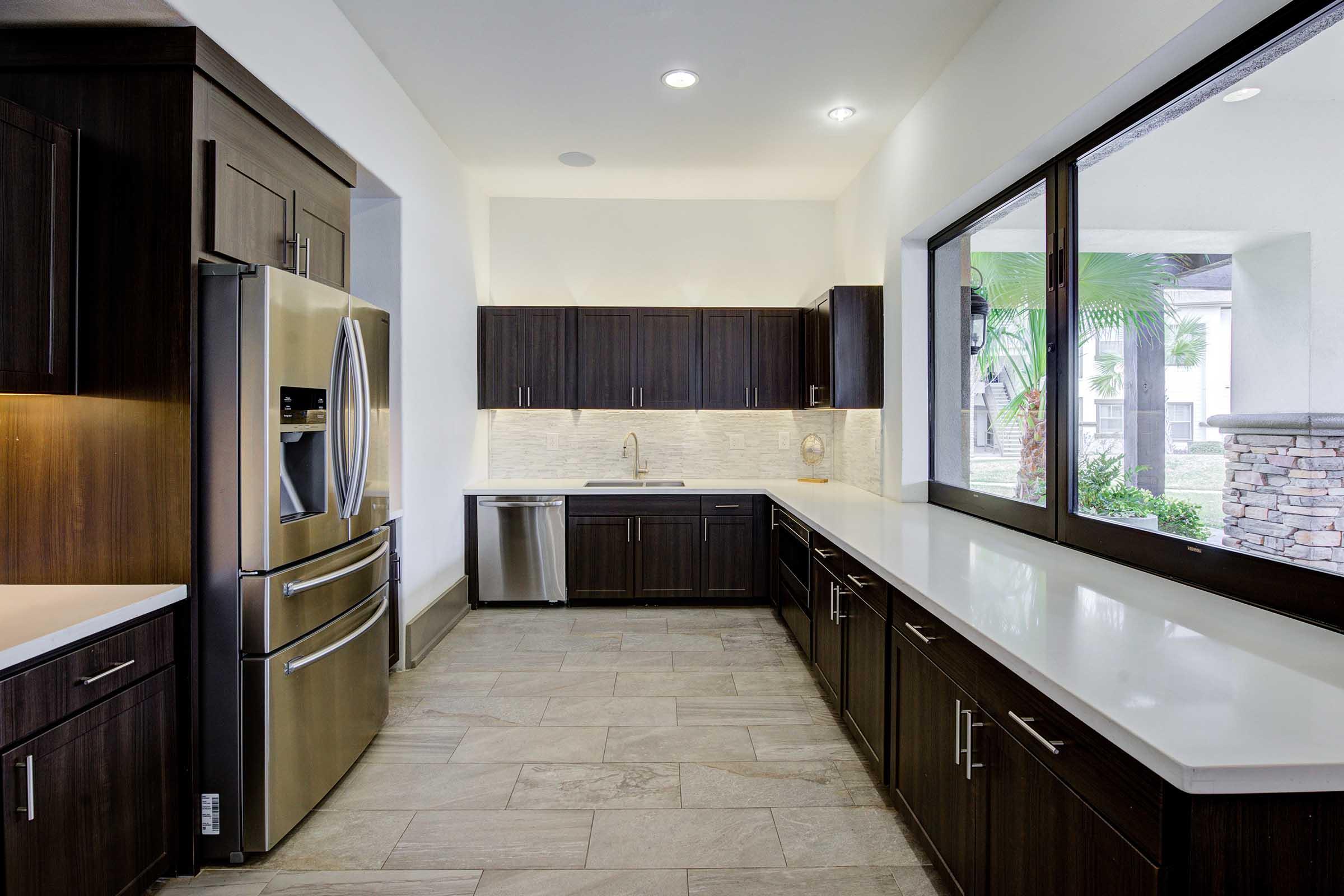 A modern kitchen featuring dark wooden cabinetry, a stainless steel refrigerator, and a sleek countertop. The space is well-lit with recessed lighting, and large windows provide a view of greenery outside. The floor is covered with textured tiles, creating a contemporary and inviting atmosphere.