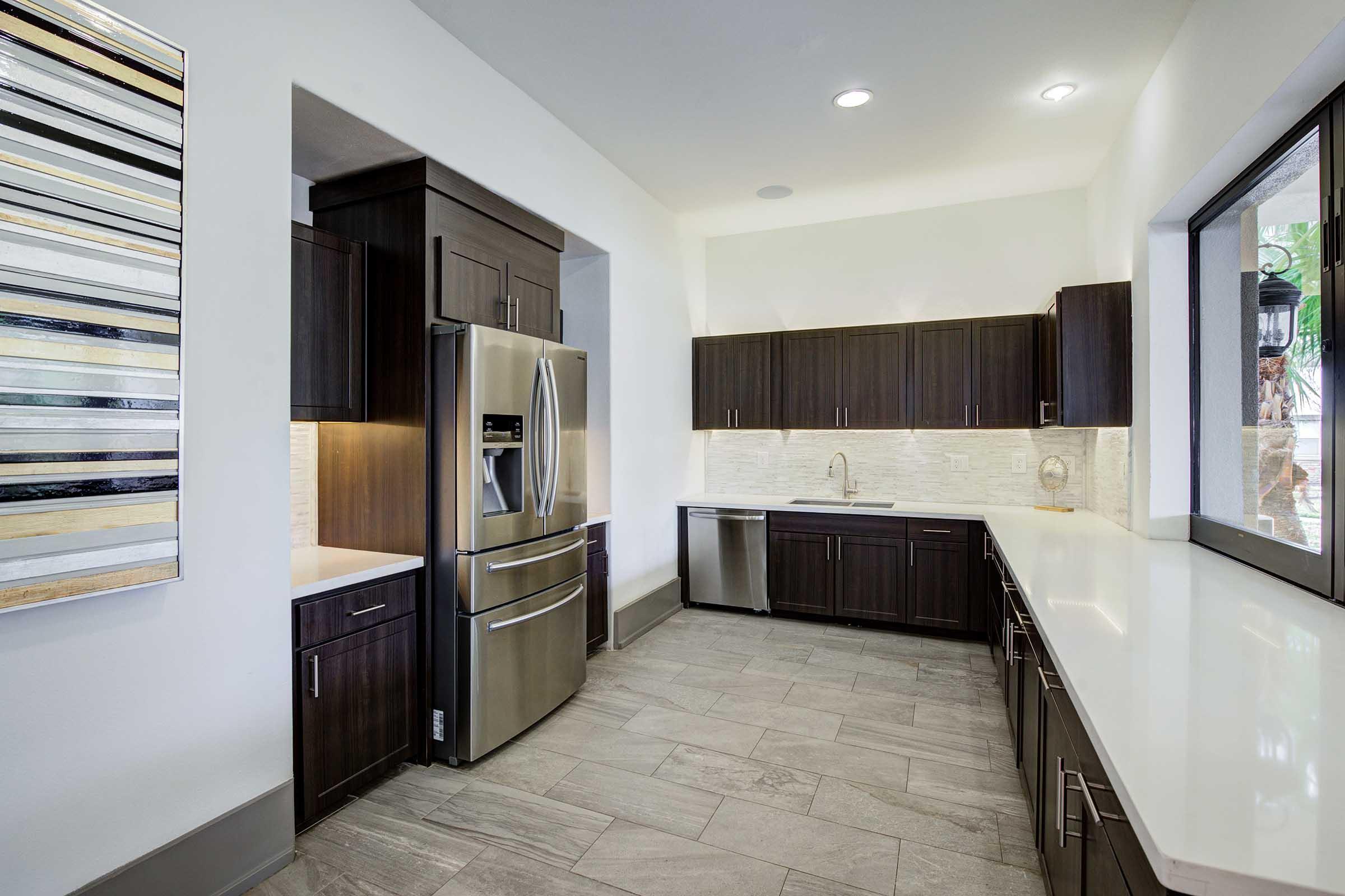 Modern kitchen featuring dark wood cabinets, stainless steel appliances, and a light-colored countertop. The space includes a refrigerator, a sink, and a dishwasher, with a sleek backsplash. Natural light comes through a window, complemented by stylish wall art. The floor is tiled with large, neutral-colored tiles.