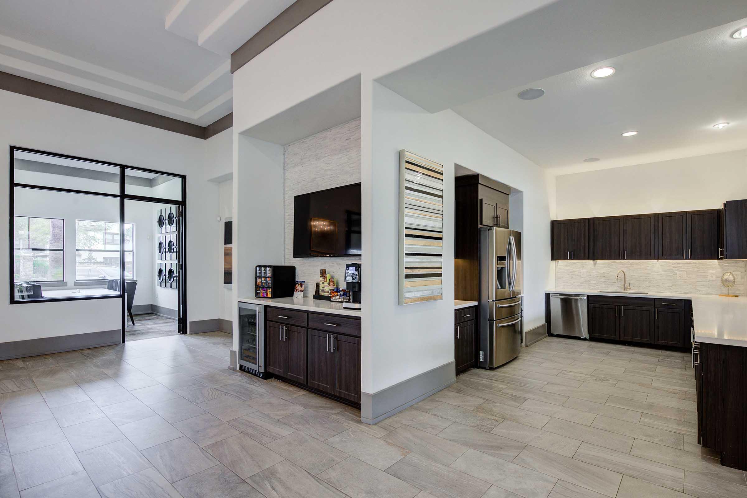 A modern, open-concept kitchen and living space featuring dark wood cabinets, stainless steel appliances, and light-colored countertops. The floor is covered with large tiles, and there is a sleek, minimalist design with ample natural light from large windows. A small dining area is visible in the background.