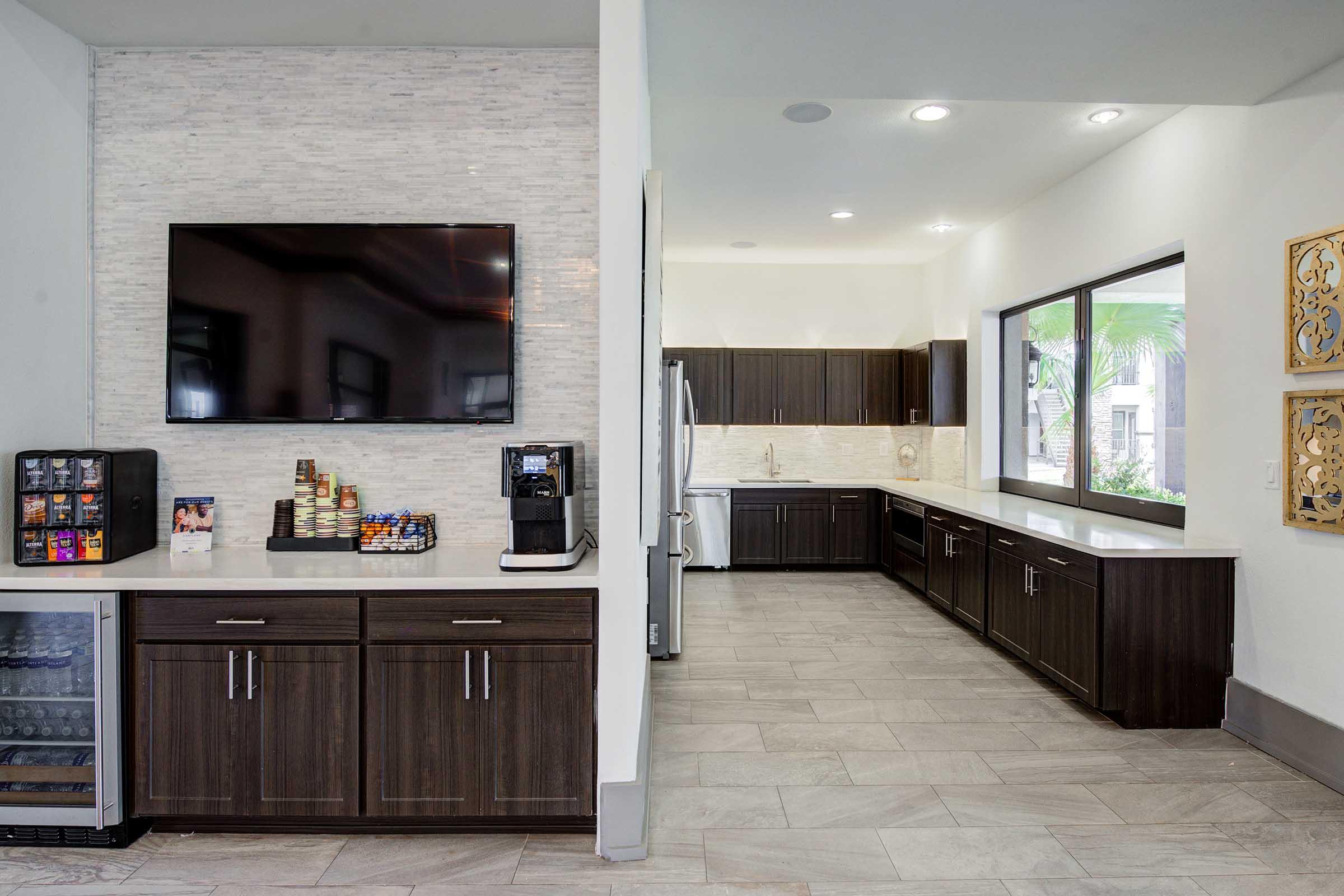 A modern kitchen space featuring dark wood cabinets, a large flat-screen TV mounted on the wall, a coffee machine, and a mini-fridge. The room has a light-colored tiled floor and large windows allowing natural light. The kitchen includes a spacious counter area with contemporary appliances.