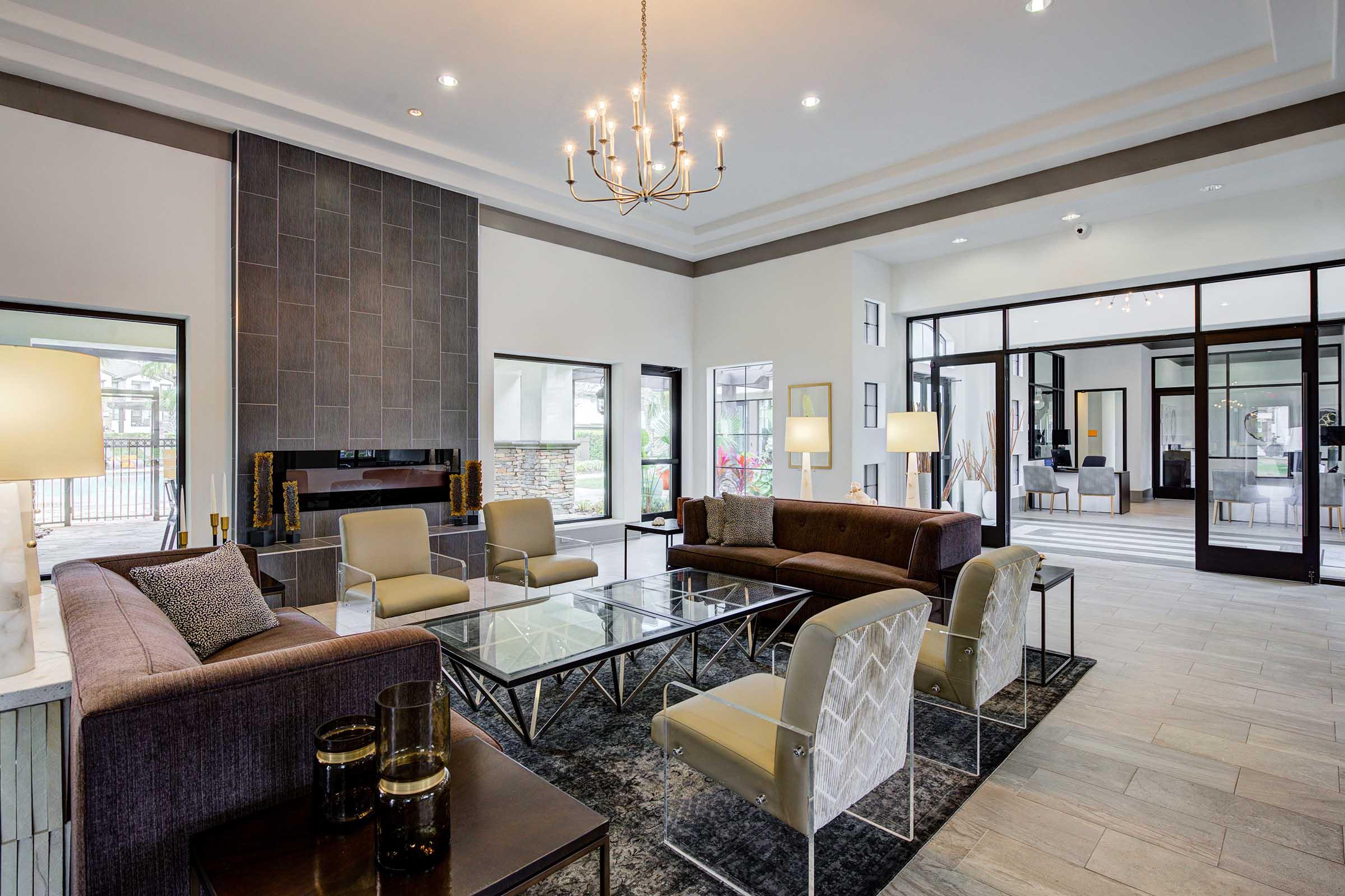 A stylish lounge area featuring modern furniture, including a dark sofa, glass coffee table, and elegant chairs. The space is brightly lit with large windows and a chandelier, showcasing a neutral color palette and contemporary decor. A cozy fireplace adds warmth to the inviting atmosphere.